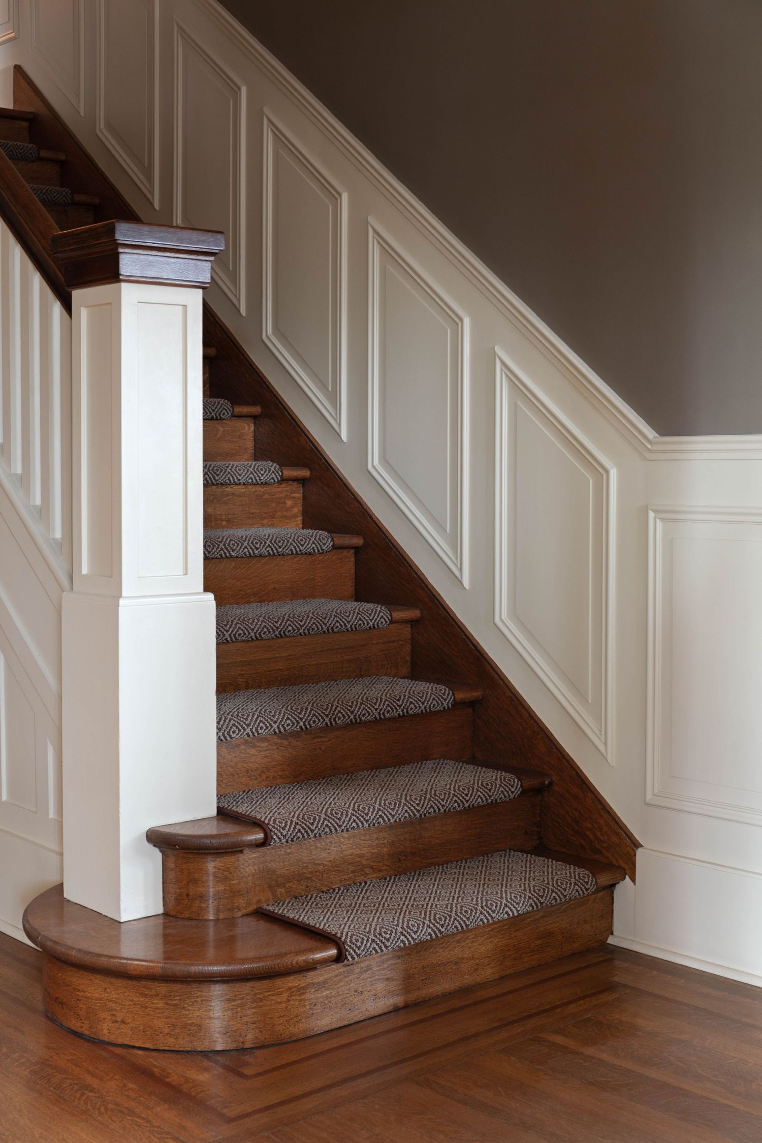 Classic wooden staircase with patterned carpet runner and white wainscoting panel walls.