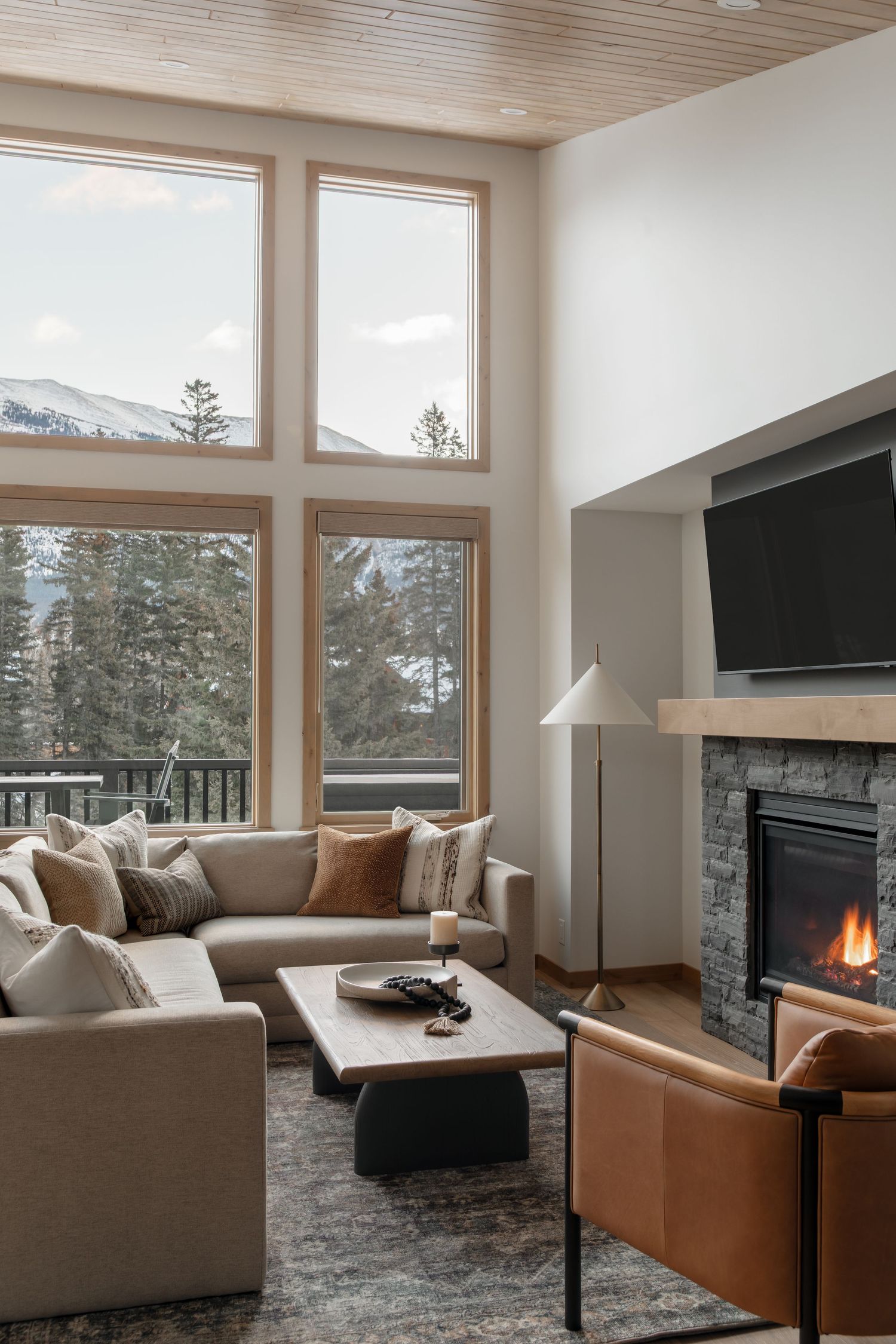 Bright mountain chalet living room with stone fireplace, sectional sofa, and floor-to-ceiling windows.