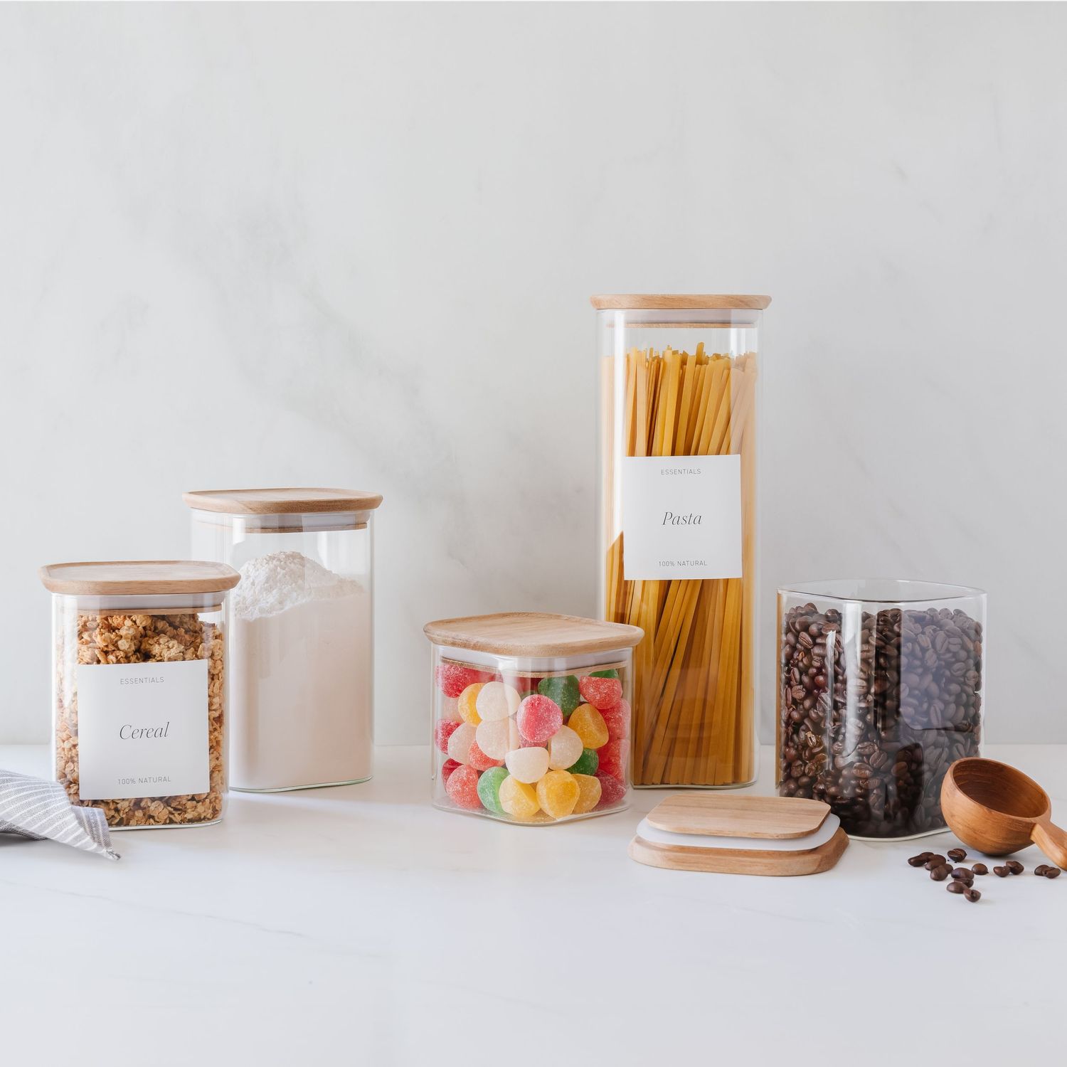 Clear glass food storage jars with bamboo lids containing pasta, cereal, candy, and coffee beans on a white surface.