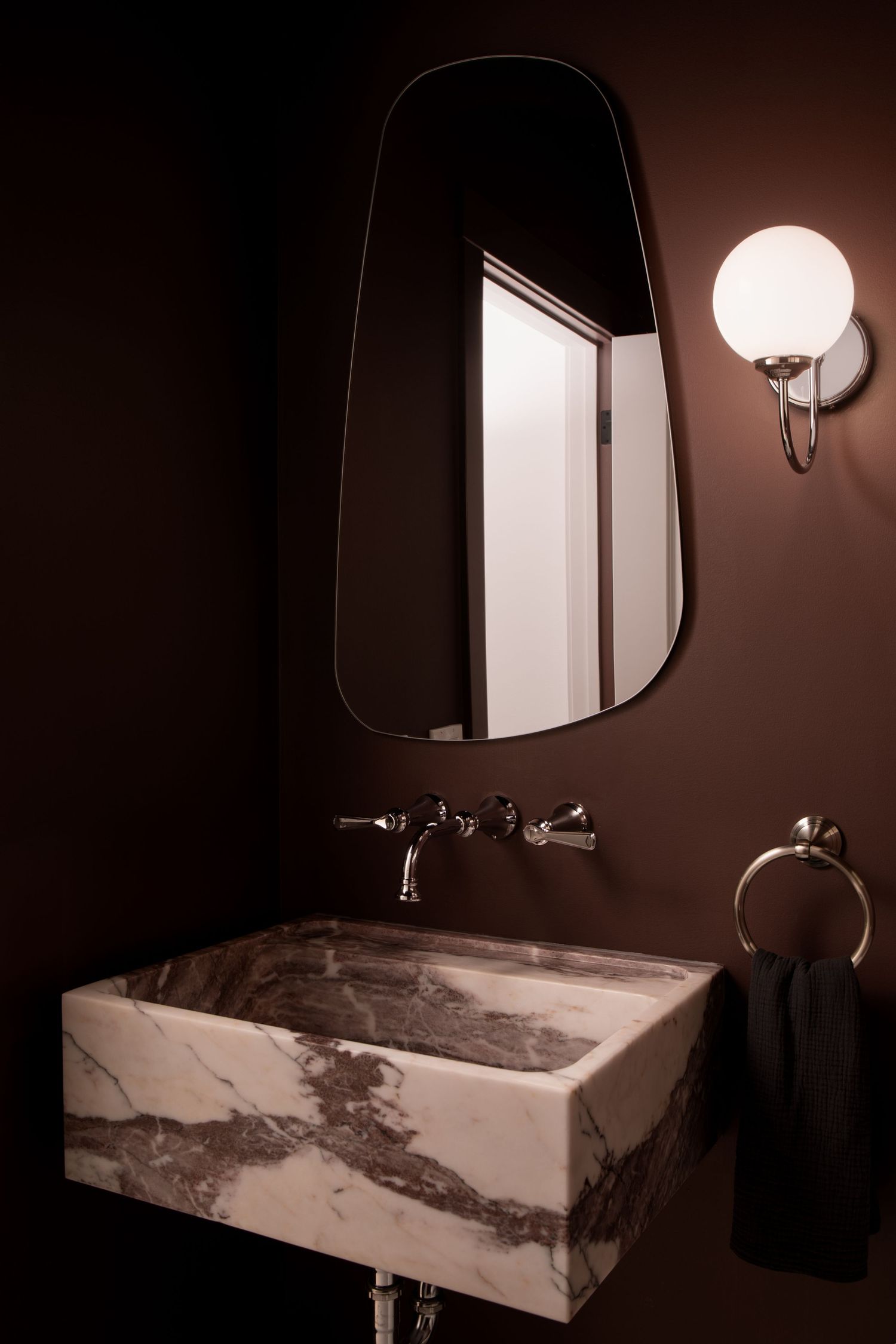 Dramatic dark powder room with marble vessel sink, globe wall sconce, and asymmetric mirror.