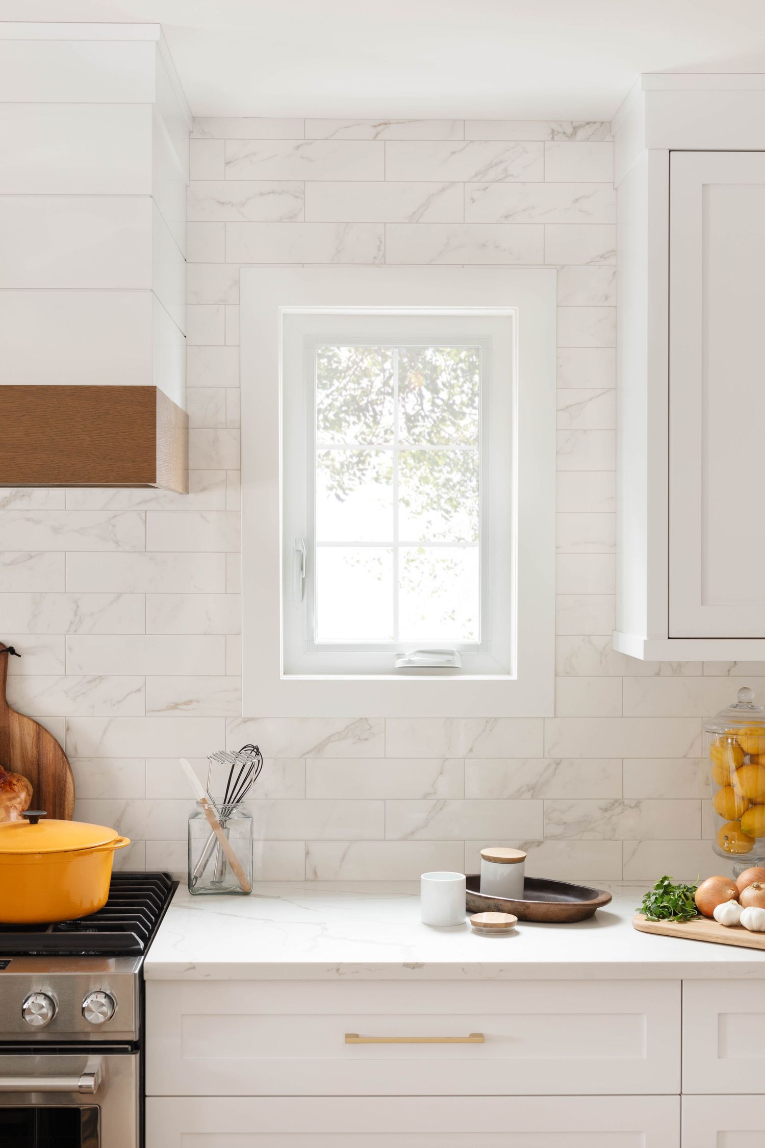 White kitchen with marble subway tile backsplash, window above counter, white cabinets, and gold hardware.