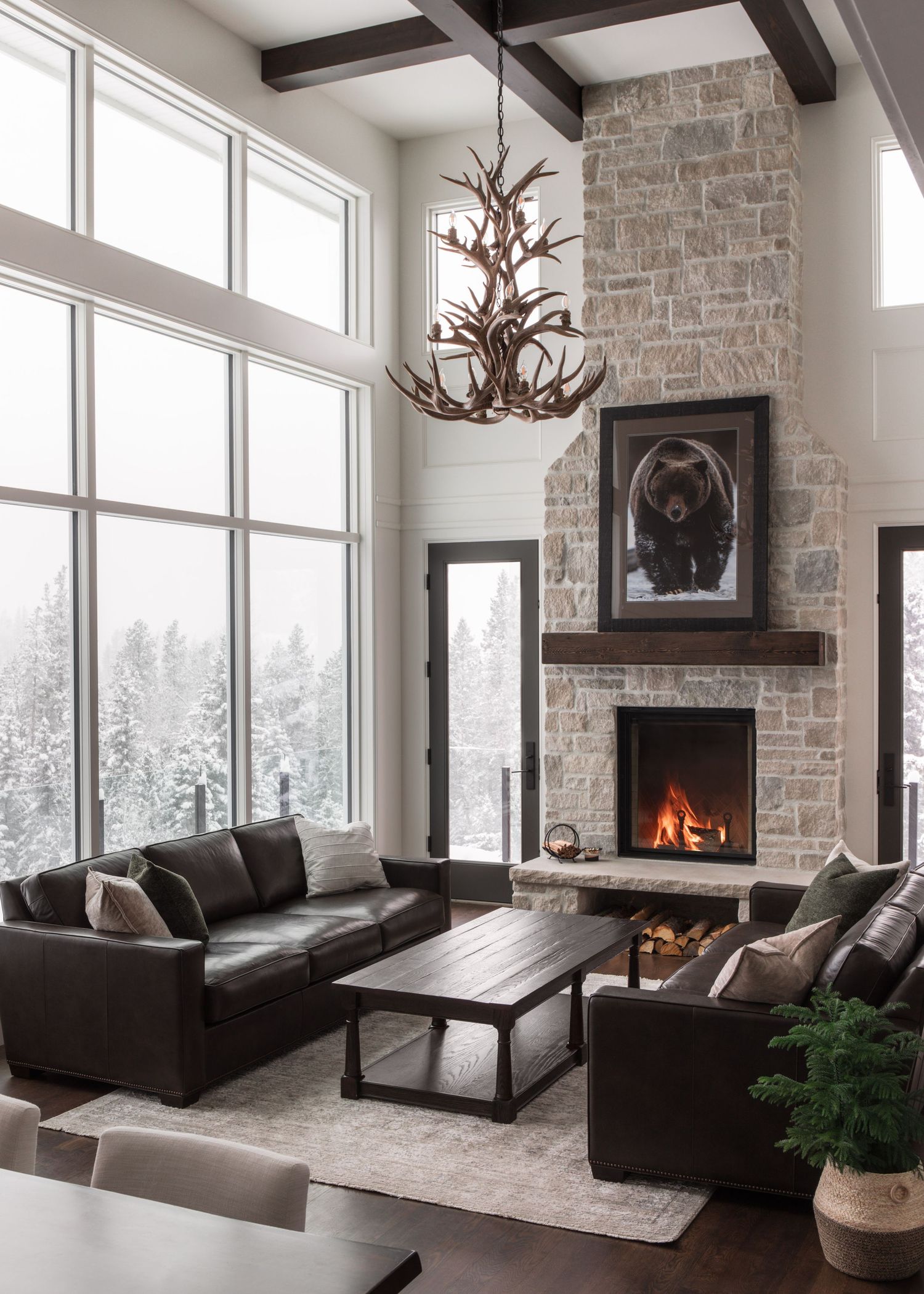 Rustic mountain living room with stone fireplace, antler chandelier, leather sofas, and floor-to-ceiling windows.