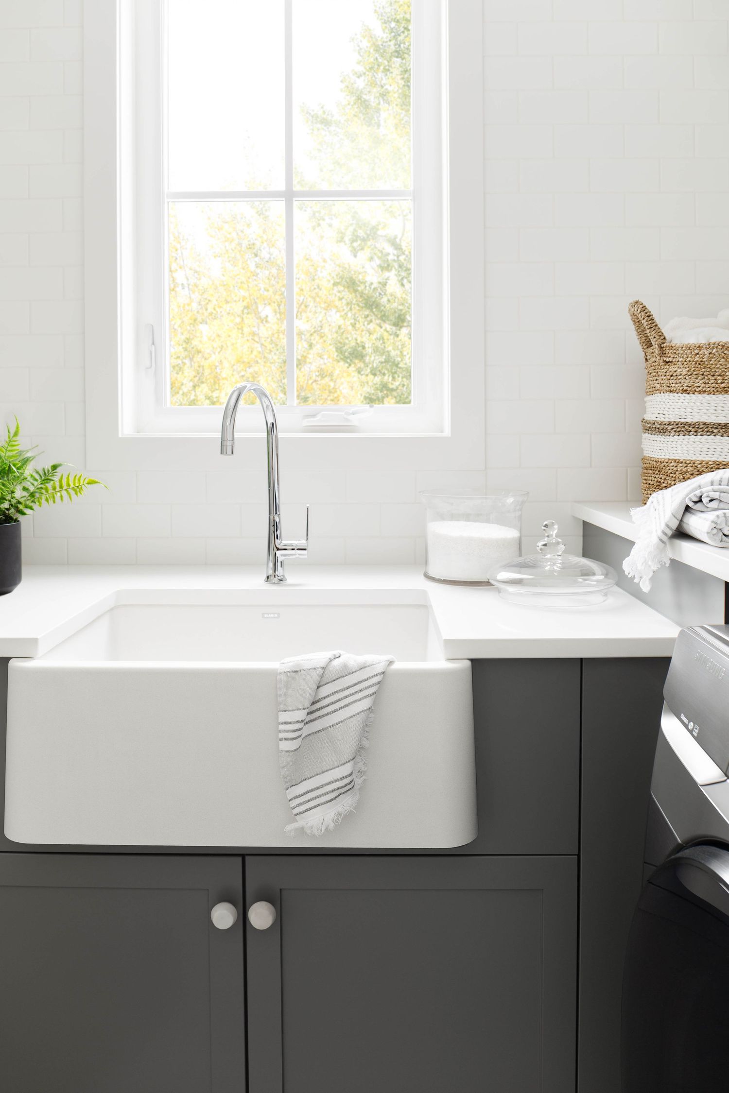 White farmhouse sink with chrome faucet in dark gray laundry room cabinet with window view.