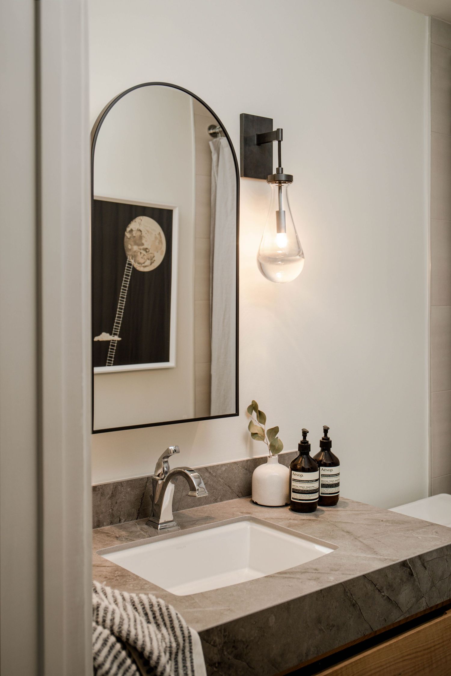 Modern bathroom vanity with marble countertop, arched mirror, black wall sconce, and Aesop products.