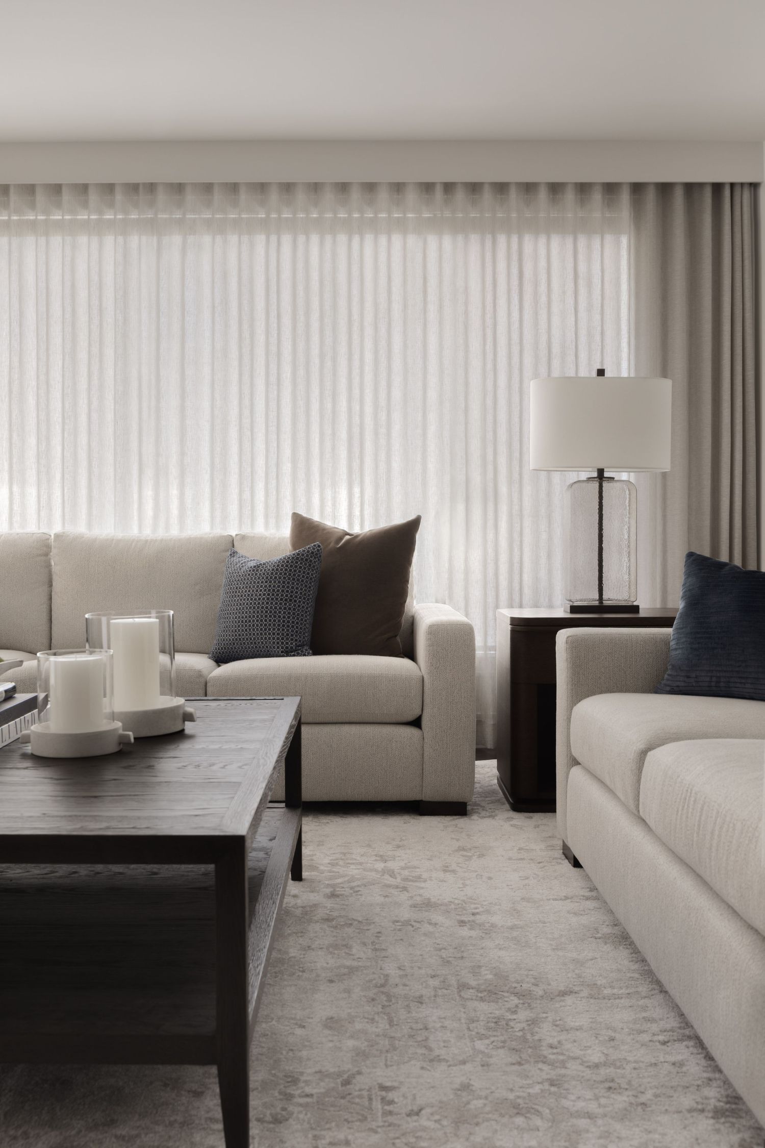 Modern neutral living room with cream sofas, dark accent pillows, table lamp, and sheer floor-to-ceiling curtains.
