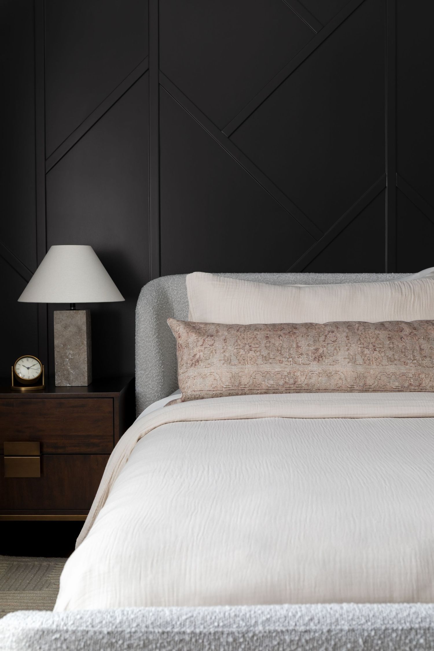Sophisticated bedroom with dark paneled wall, gray upholstered bed, white bedding, and marble nightstand lamp.