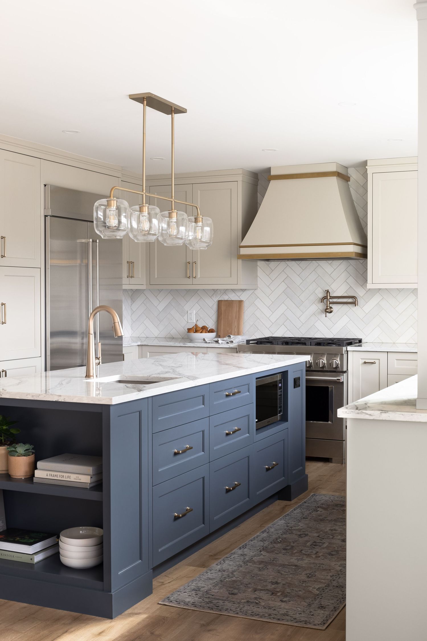 Traditional kitchen with navy blue island, brass pendant lights, herringbone tile backsplash, and custom range hood.