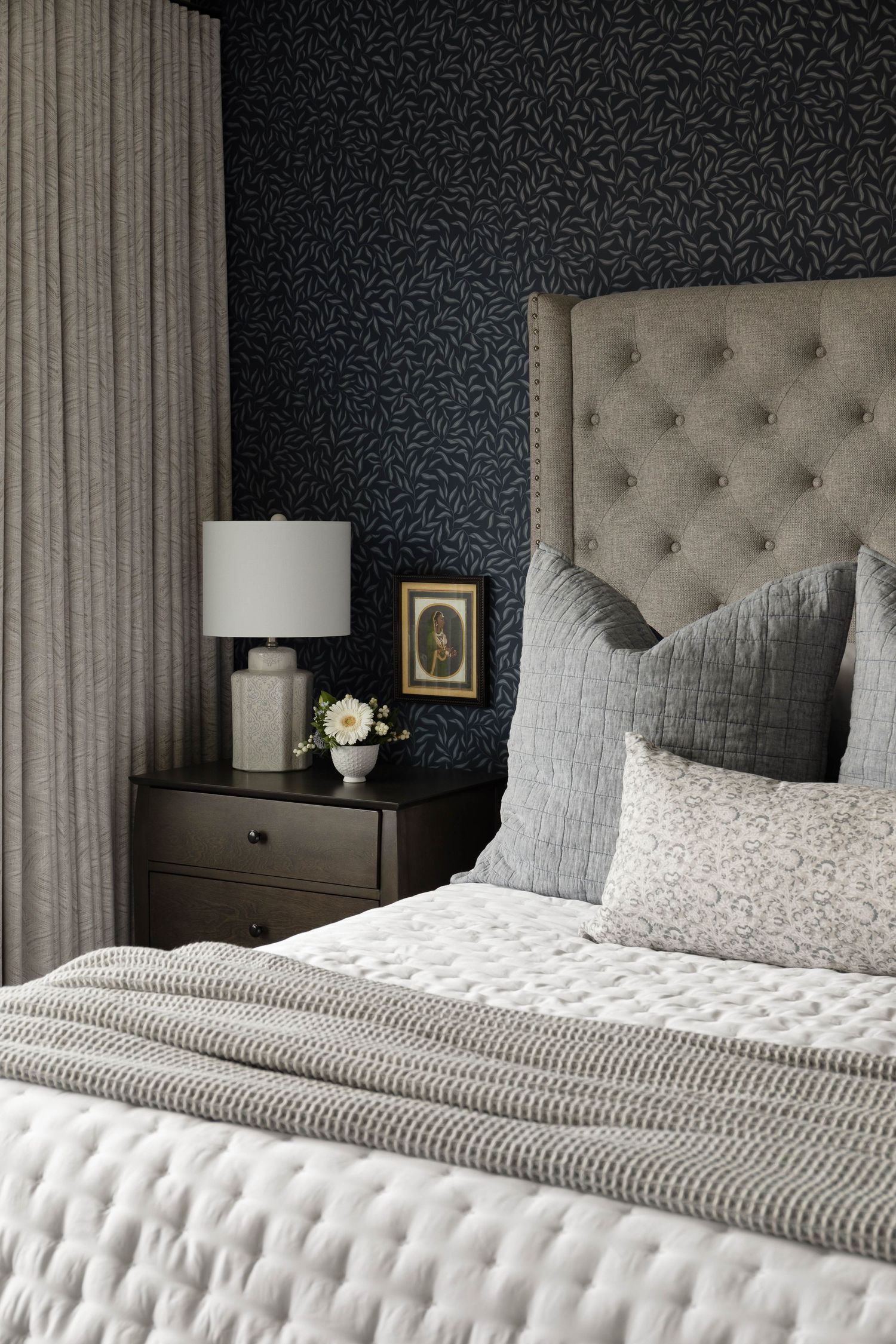 Elegant bedroom with tufted gray headboard, layered neutral bedding, dark nightstand, and navy botanical wallpaper.