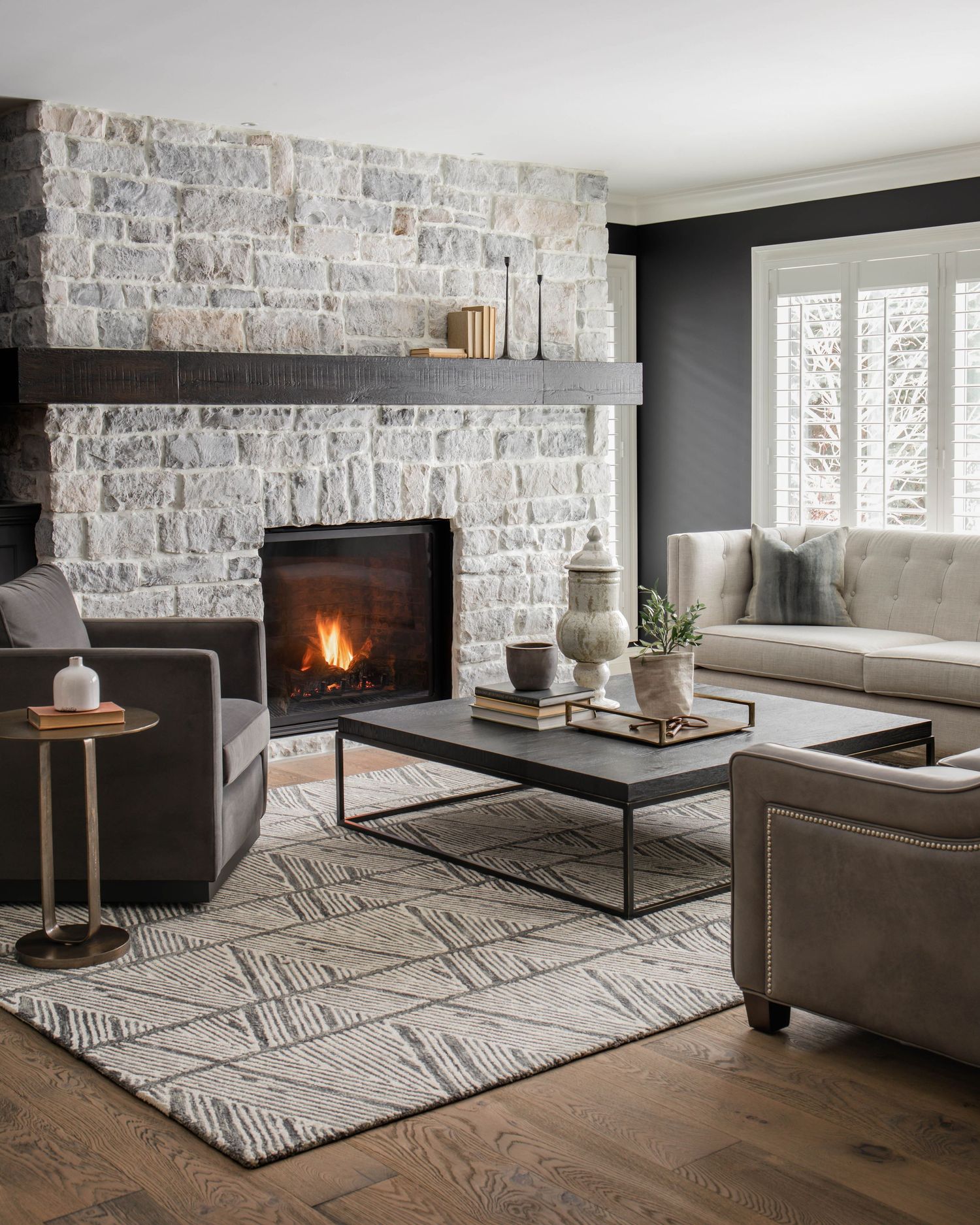 Cozy living room with stone fireplace, dark walls, gray leather sofas, geometric area rug, and lit fire.
