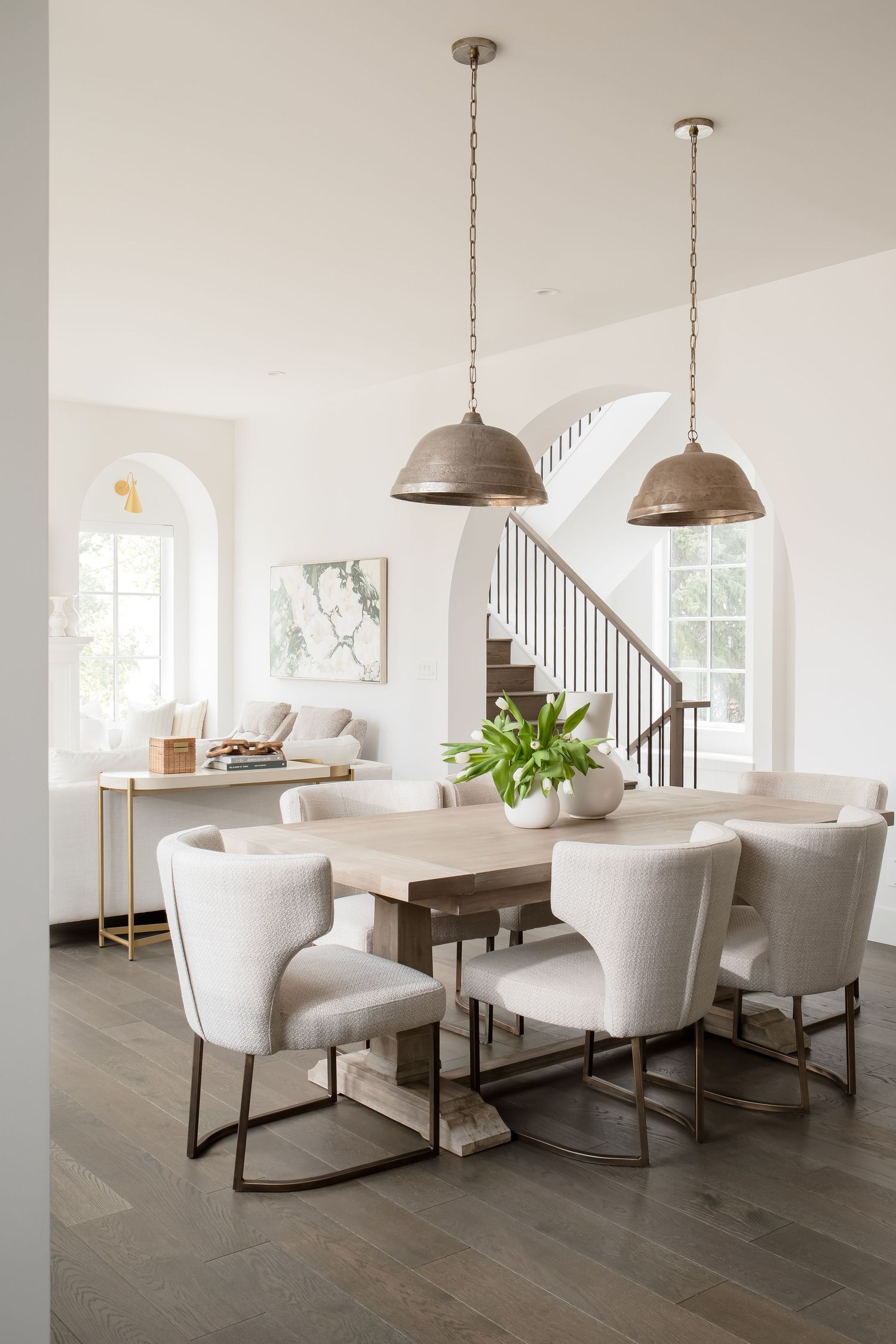 Airy dining room with light wood table, upholstered chairs, bronze dome pendants, and arched windows.