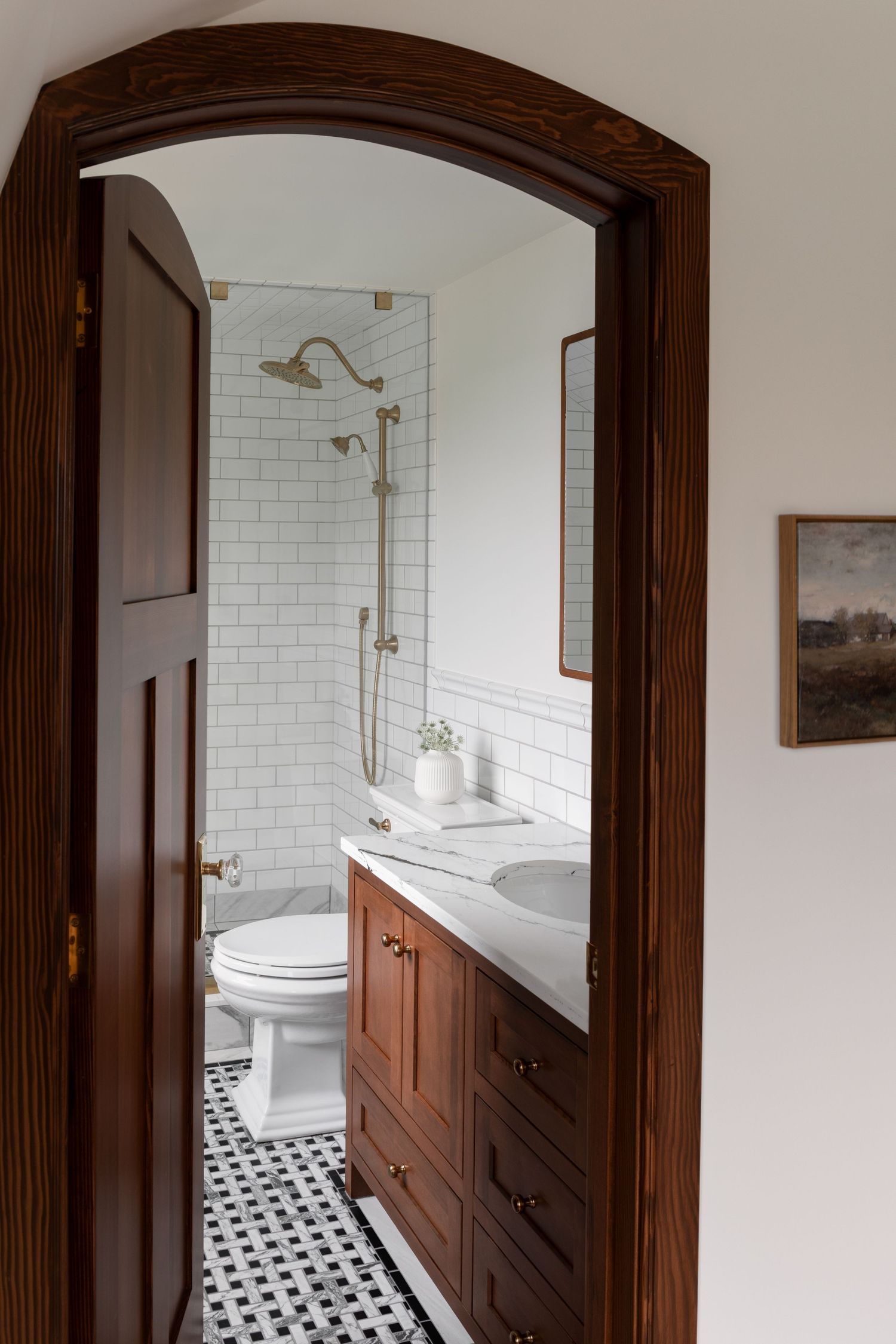 Elegant bathroom with dark wood vanity, white subway tiles, brass fixtures, black and white mosaic floor, and arched doorway.