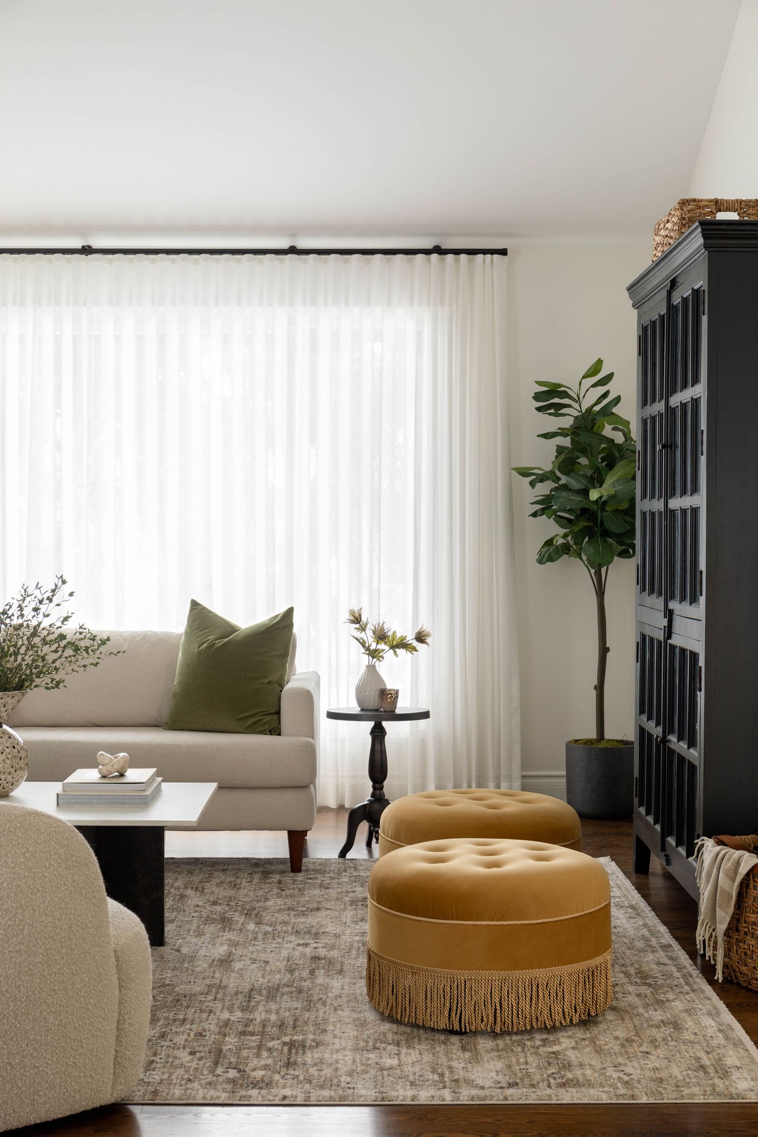 Bright living room featuring a beige sofa, mustard tufted ottoman, black cabinet, and fiddle leaf fig tree.