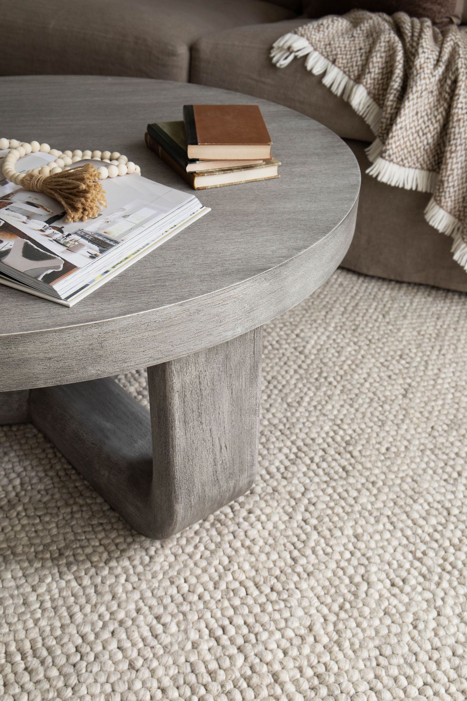 Round gray wood coffee table with books and beaded garland on neutral woven rug.