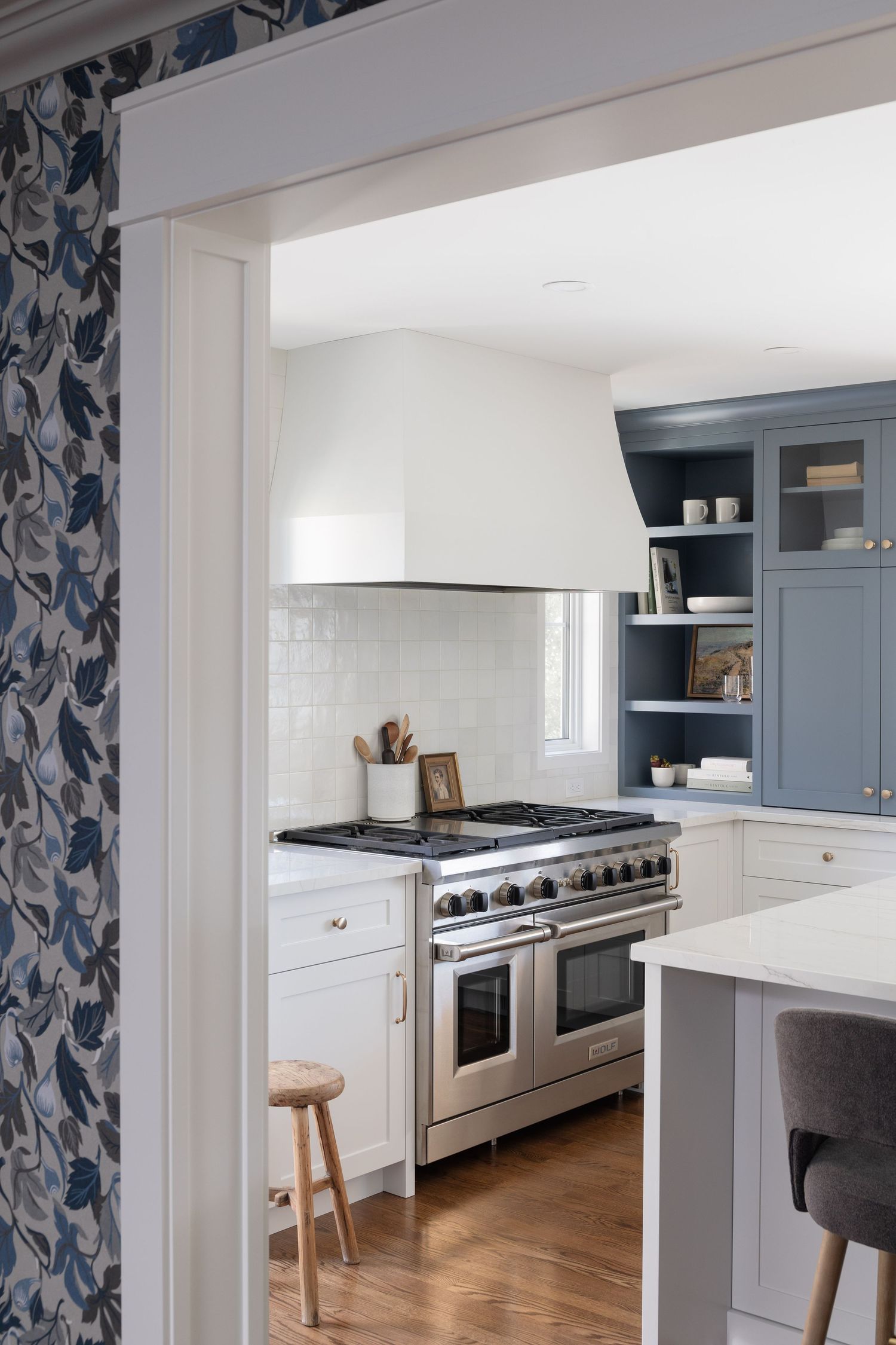 Blue-gray kitchen cabinets with open shelving, professional stainless range, white hood, and floral wallpaper doorway.