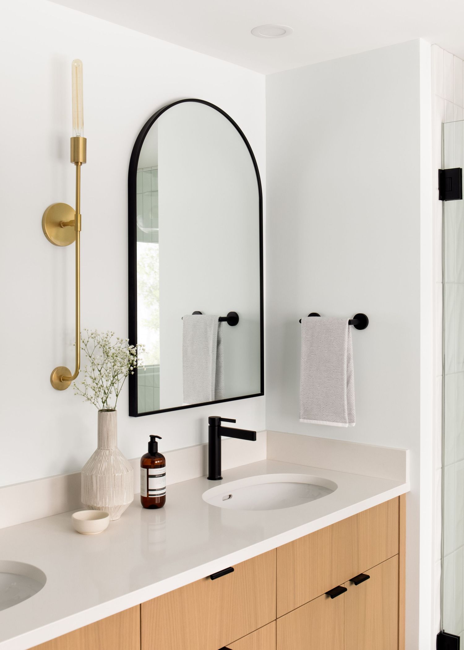 Modern bathroom vanity with arched black mirror, brass sconce, wood cabinets, and matte black fixtures.