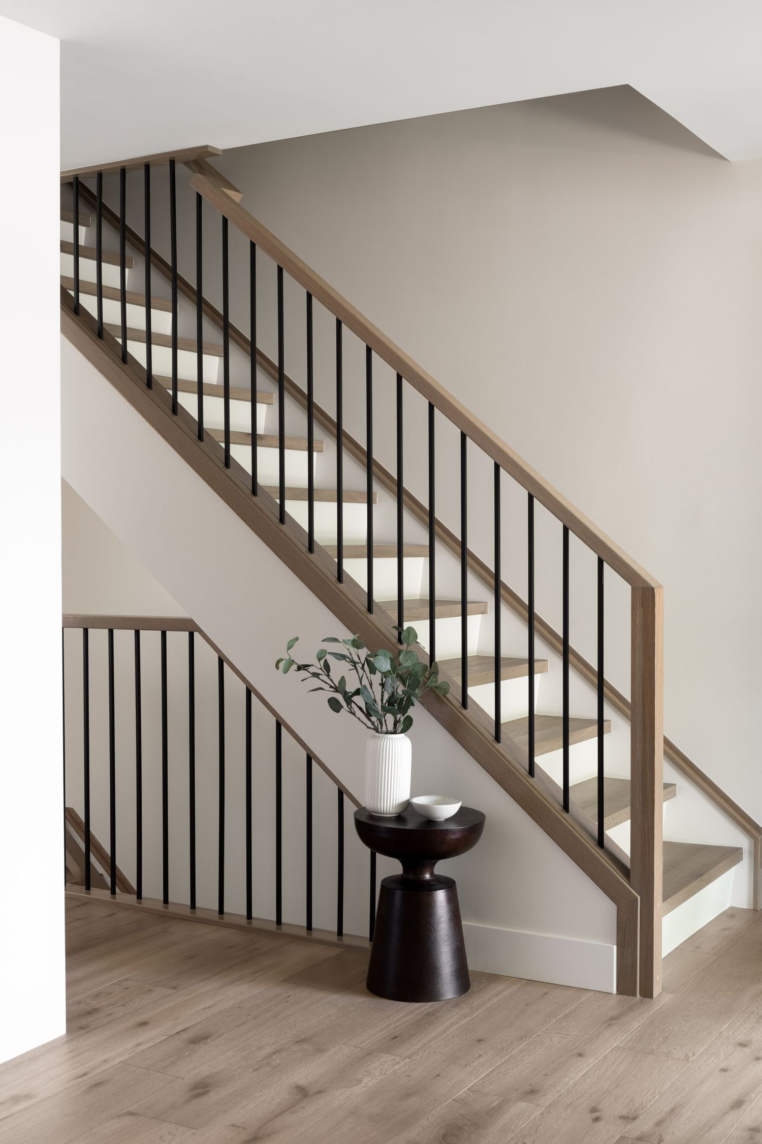 Modern staircase with warm wood handrail, black metal balusters, white risers, and a dark wood side table with plant decor.