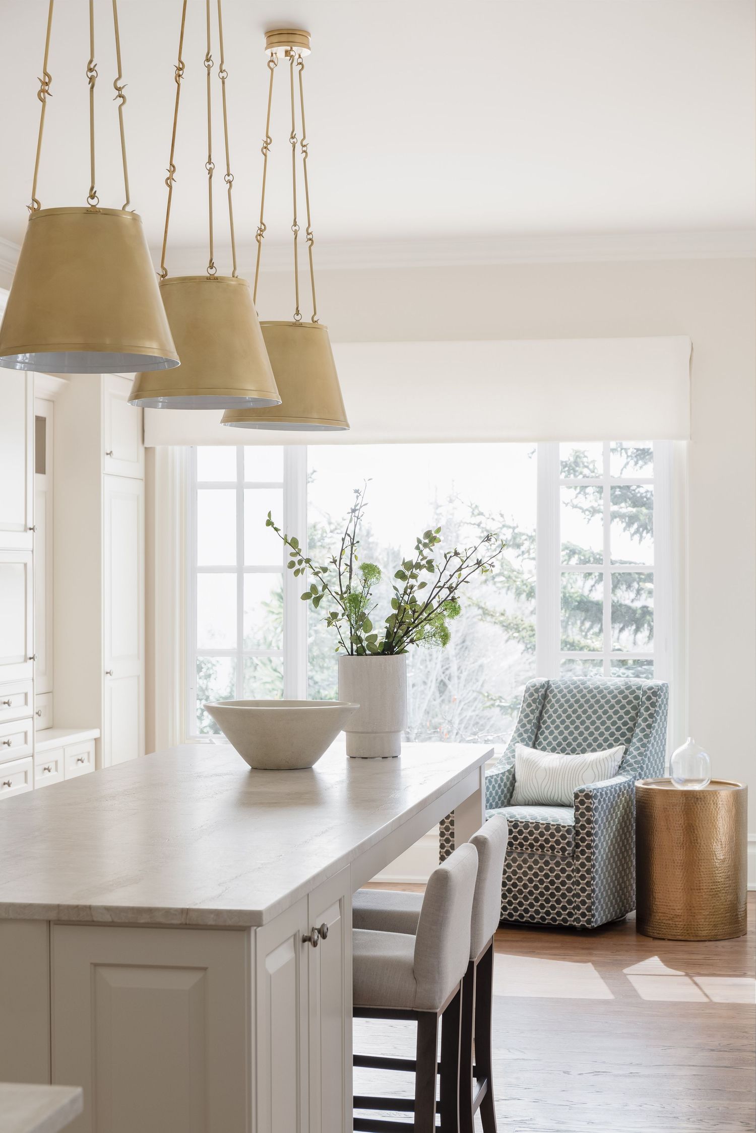 Bright kitchen with gold pendant lights, white island, upholstered barstools, and garden window.