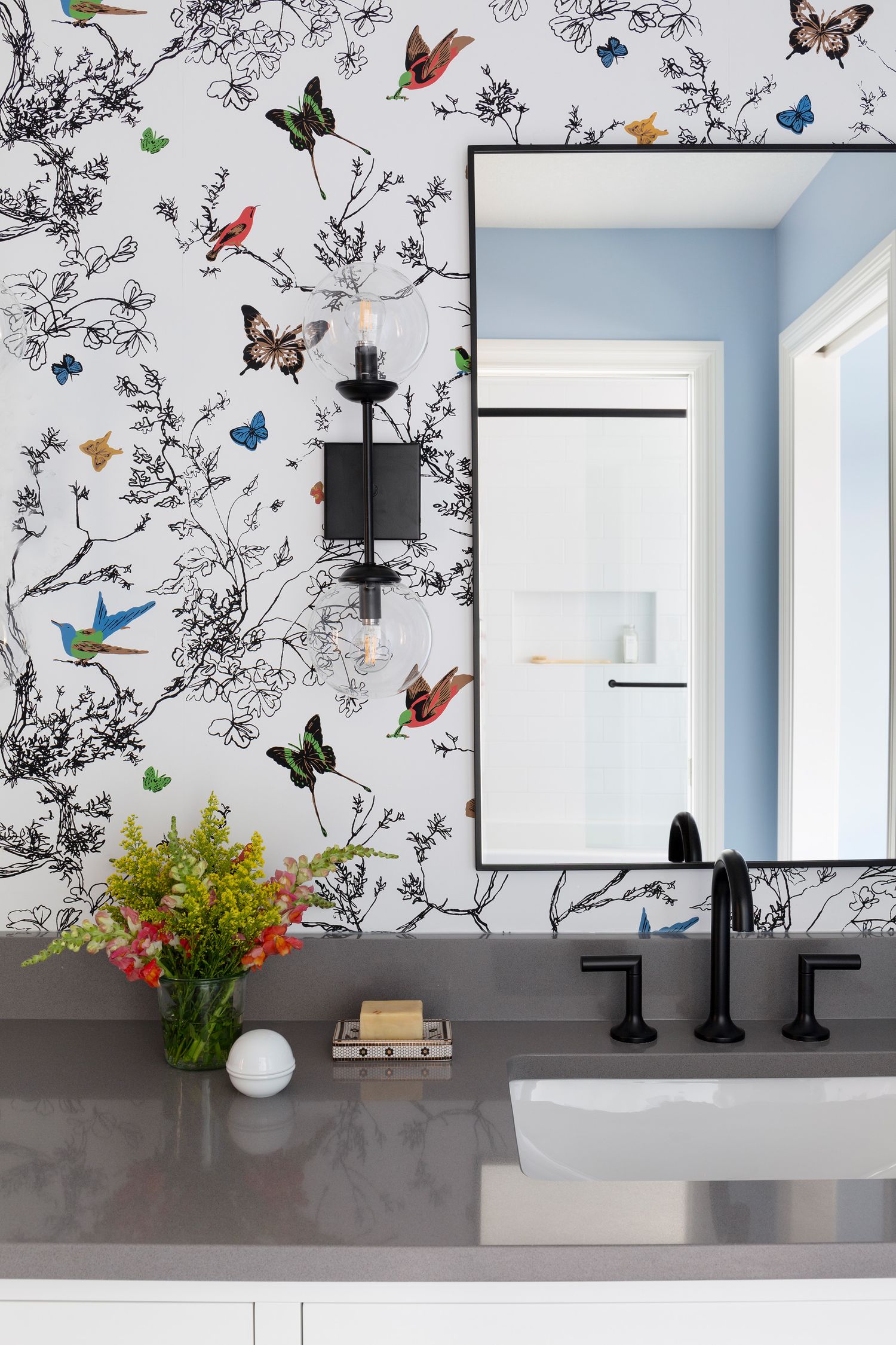 Bathroom vanity with butterfly wallpaper, black fixtures, gray countertop, and rectangular mirror.