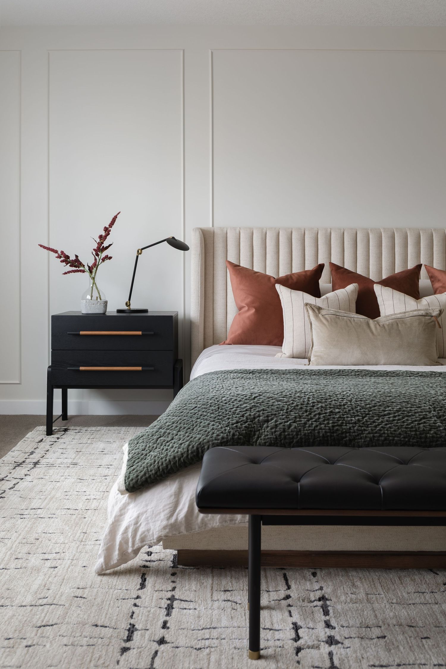 Elegant bedroom with beige tufted headboard, terracotta pillows, green throw, black nightstand, and patterned rug.