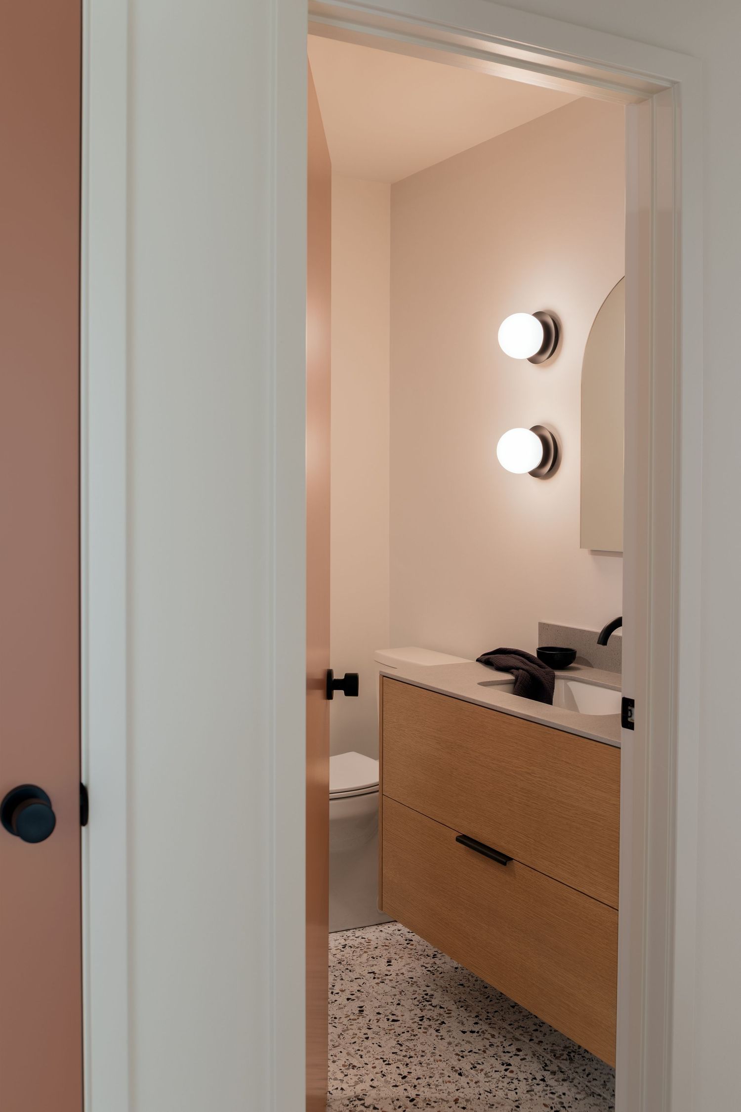 Compact powder room with warm pink walls, floating wood vanity, terrazzo floor, and round globe wall sconces.