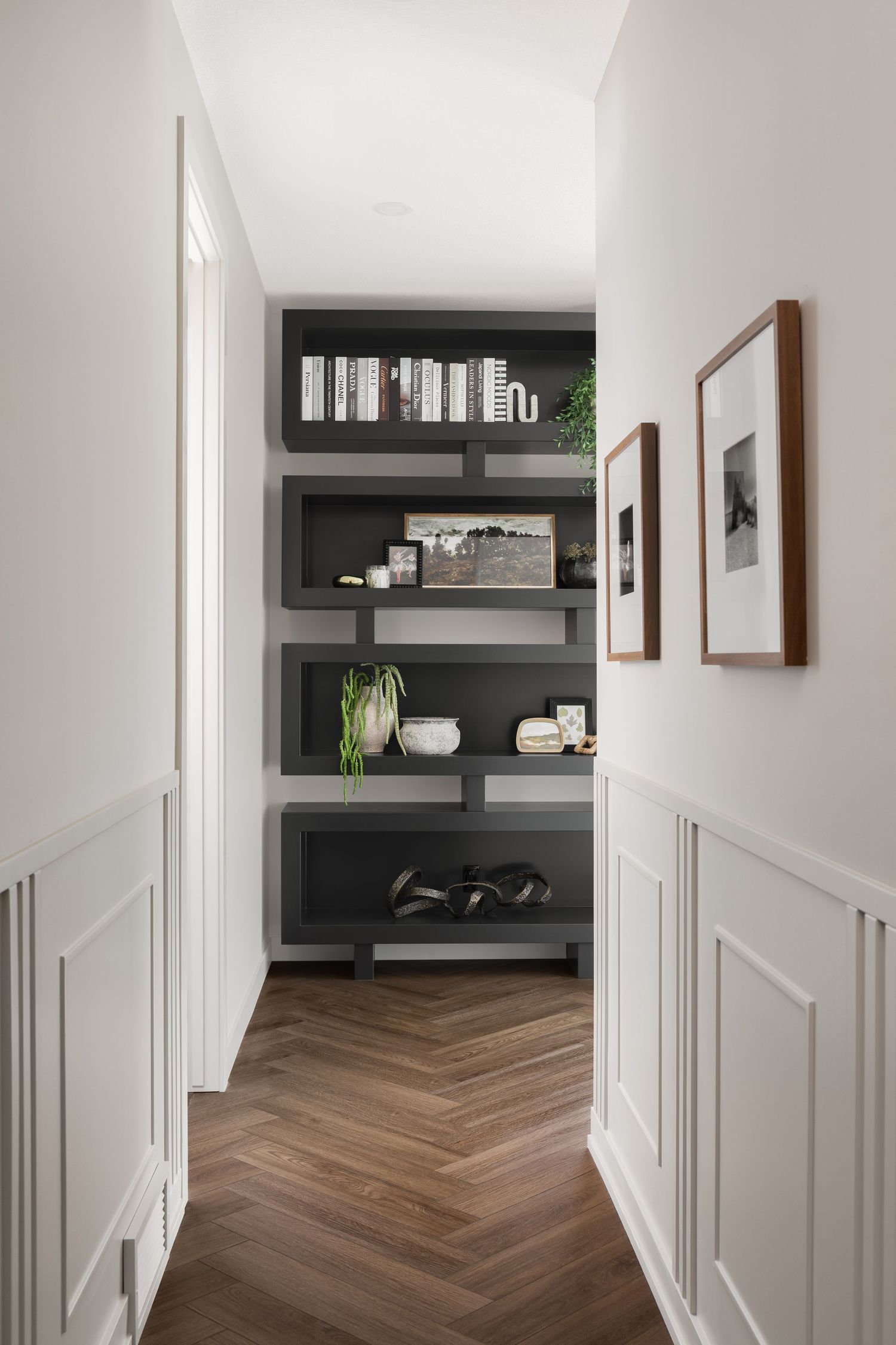 Elegant hallway with herringbone wood floors, white wainscoting, and dark built-in shelving with decor.