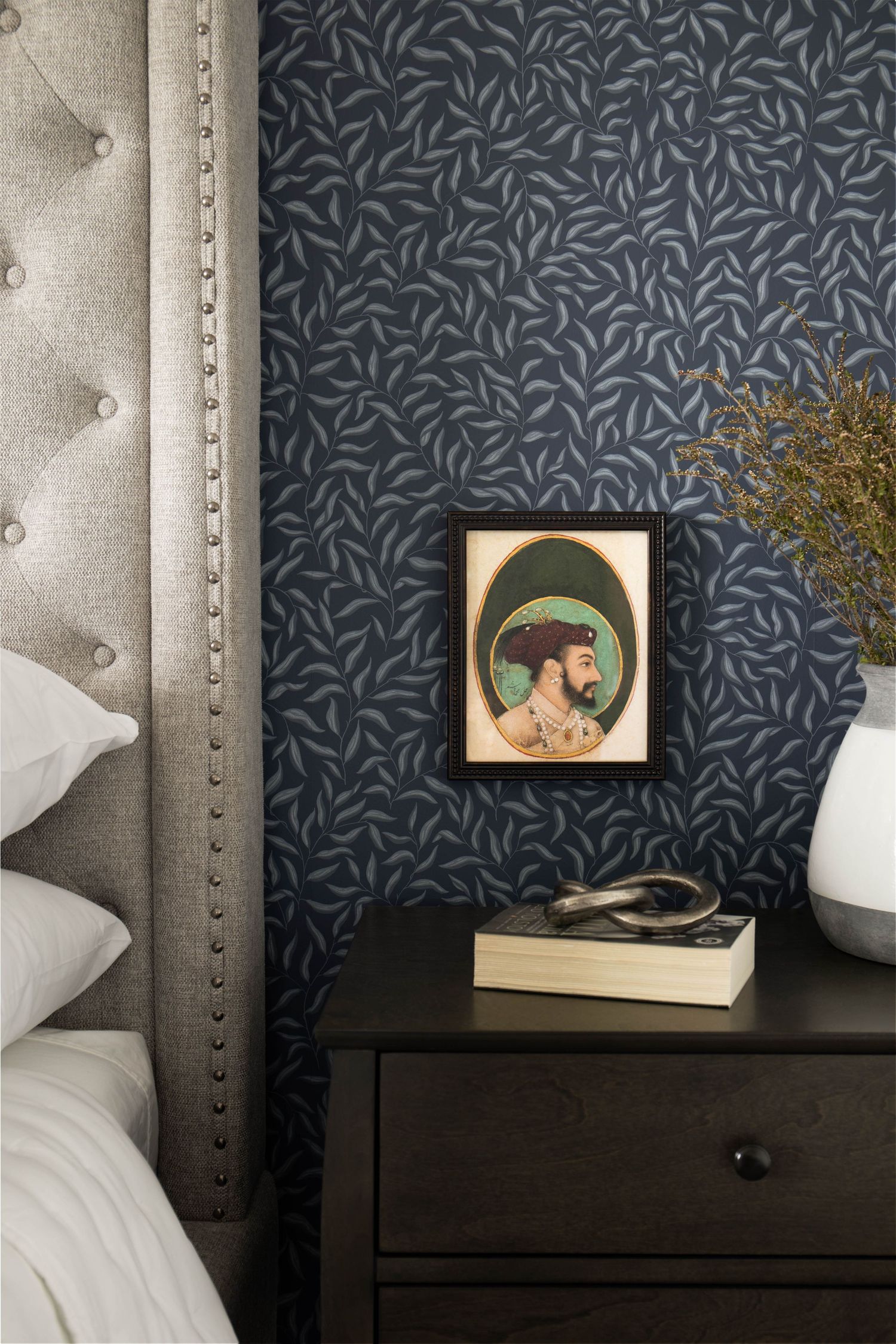 Dark nightstand with framed portrait art and books against navy leaf-patterned wallpaper beside tufted headboard.