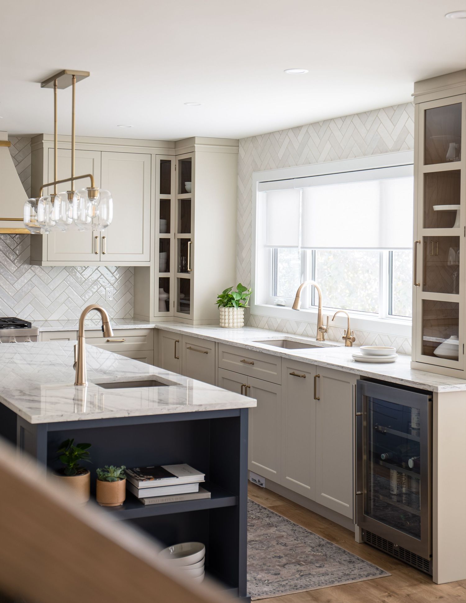 Modern kitchen with cream cabinets, navy island, gold fixtures, herringbone backsplash, quartz countertops, and wine fridge near window.