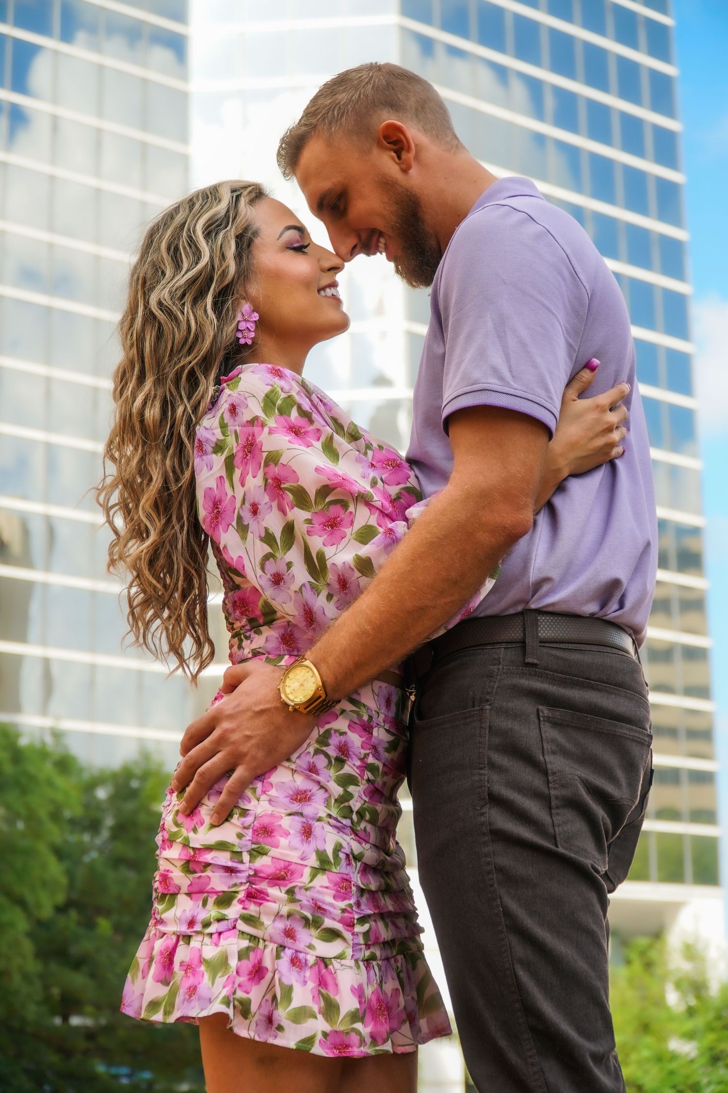 Romantic couple embraces outdoors with a floral dress and purple shirt against modern glass building backdrop.
