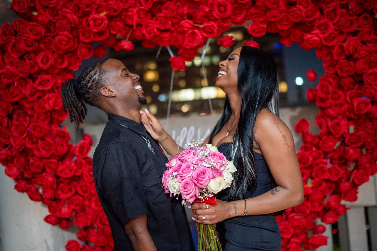 A romantic couple sharing laughter near a heart-shaped red rose wall with pink bouquet.