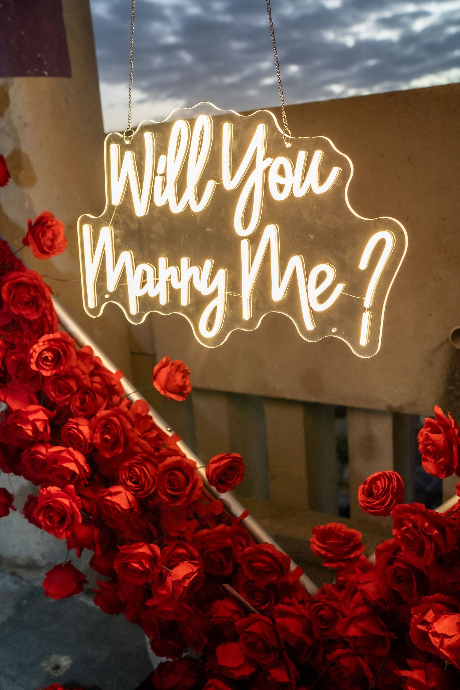 A glowing neon sign reading 'Will You Marry Me?' surrounded by vibrant red roses against a moody evening sky.