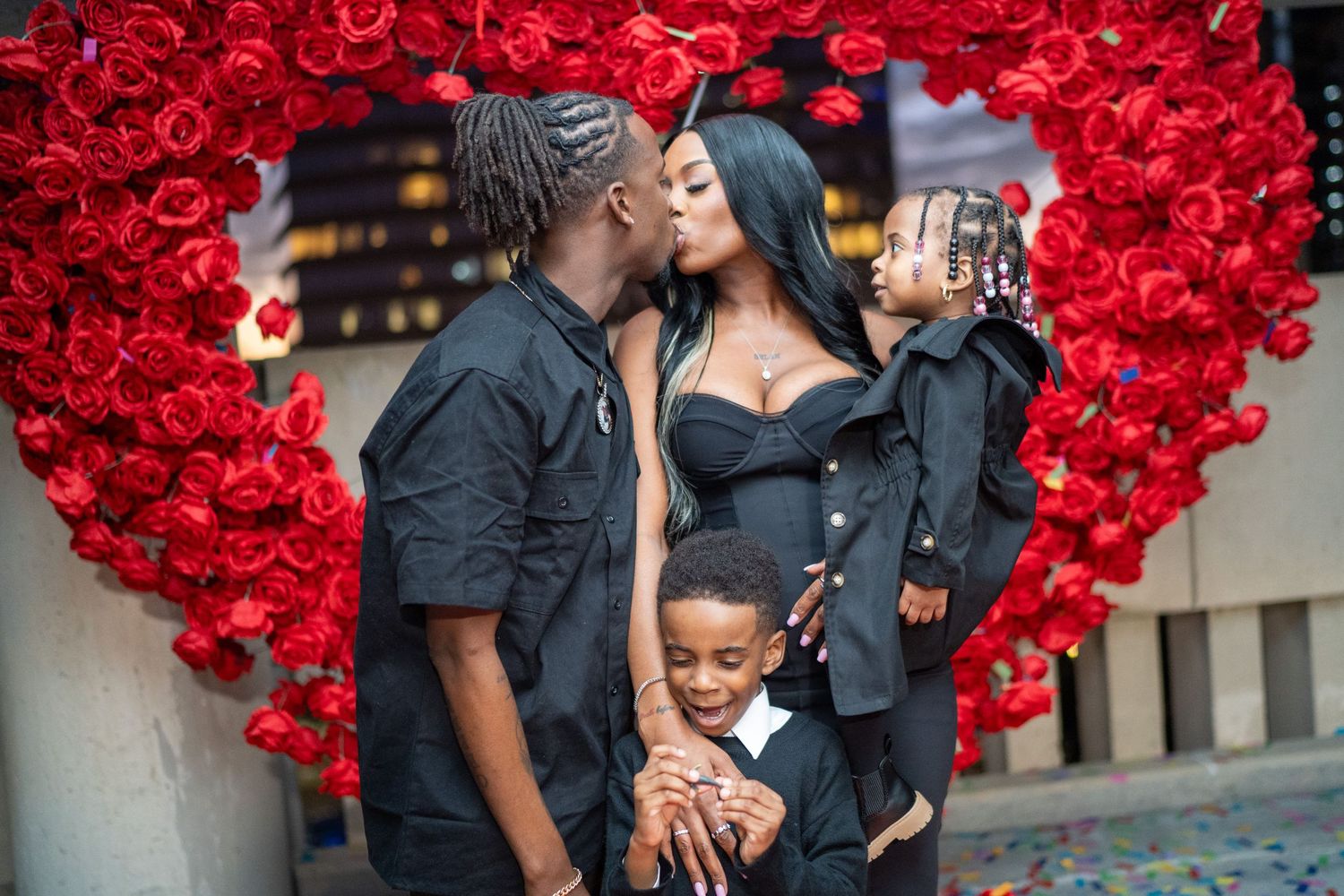 A family shares a tender moment in front of a large heart-shaped display made of vibrant red roses.