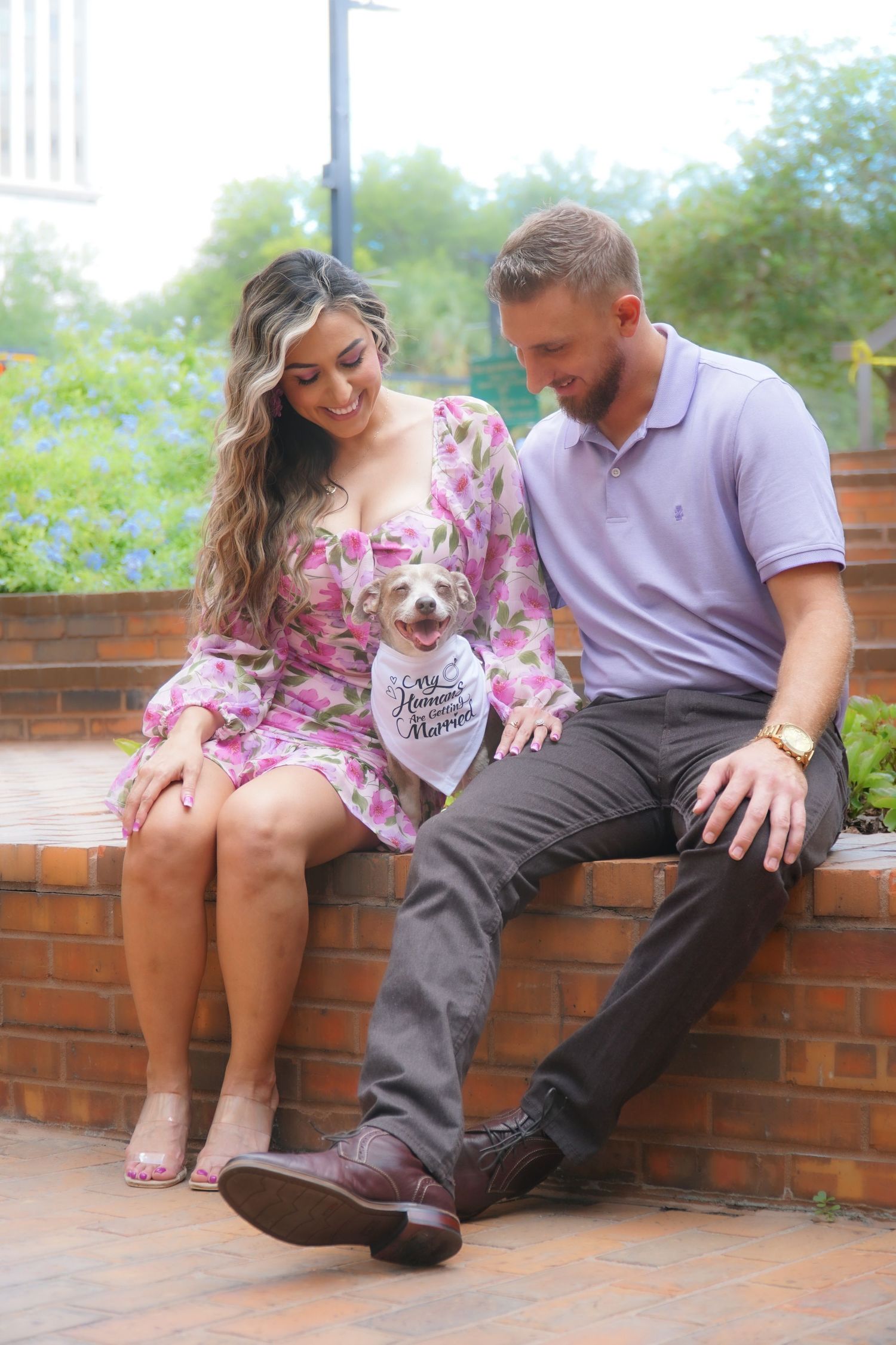 A couple sits on a brick ledge outdoors with their small dog wearing a bandana amid greenery and trees.