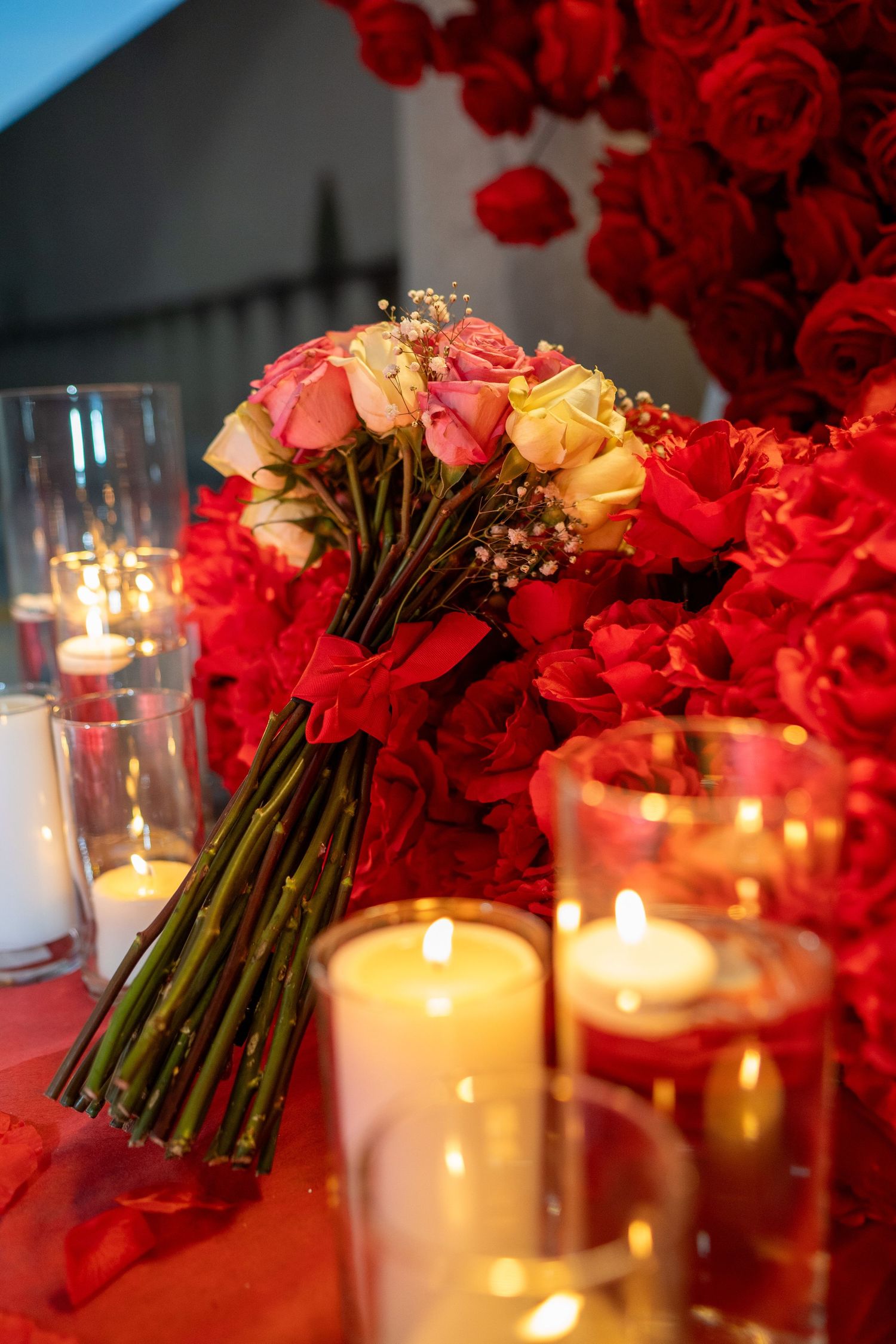 A romantic arrangement of red and coral roses with baby's breath sits beside glowing white candles on a red tablecloth.