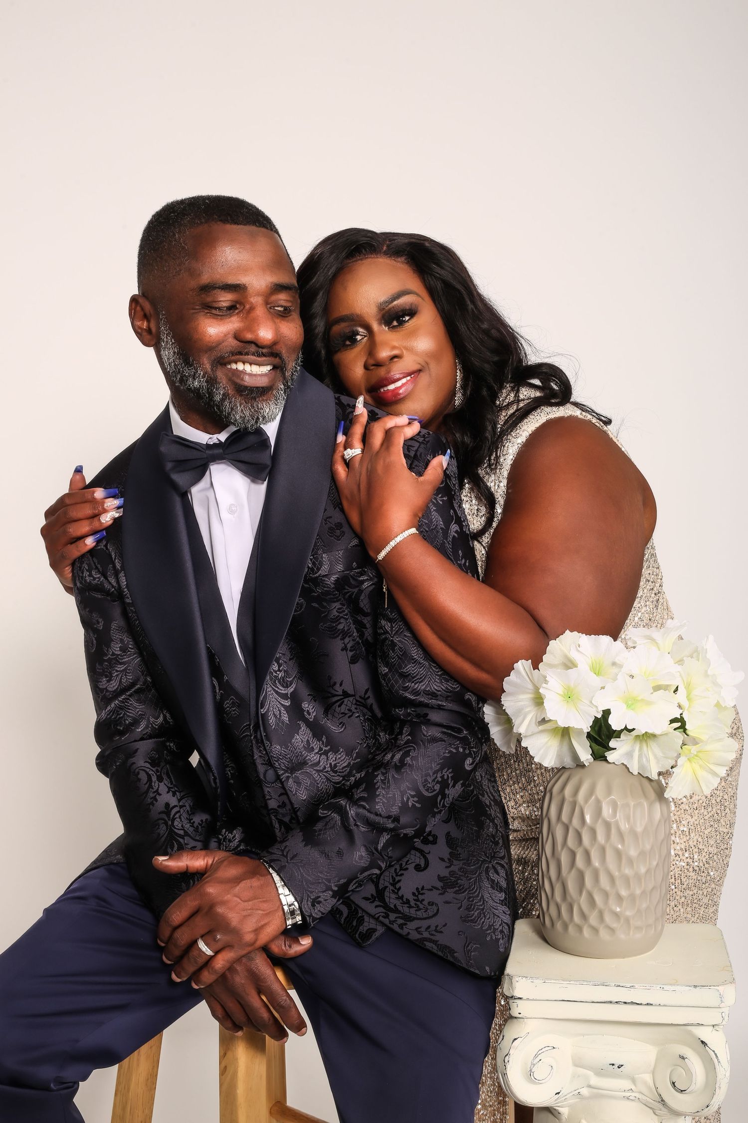 A couple in elegant formal wear pose together near white flowers in a studio setting with a neutral background.