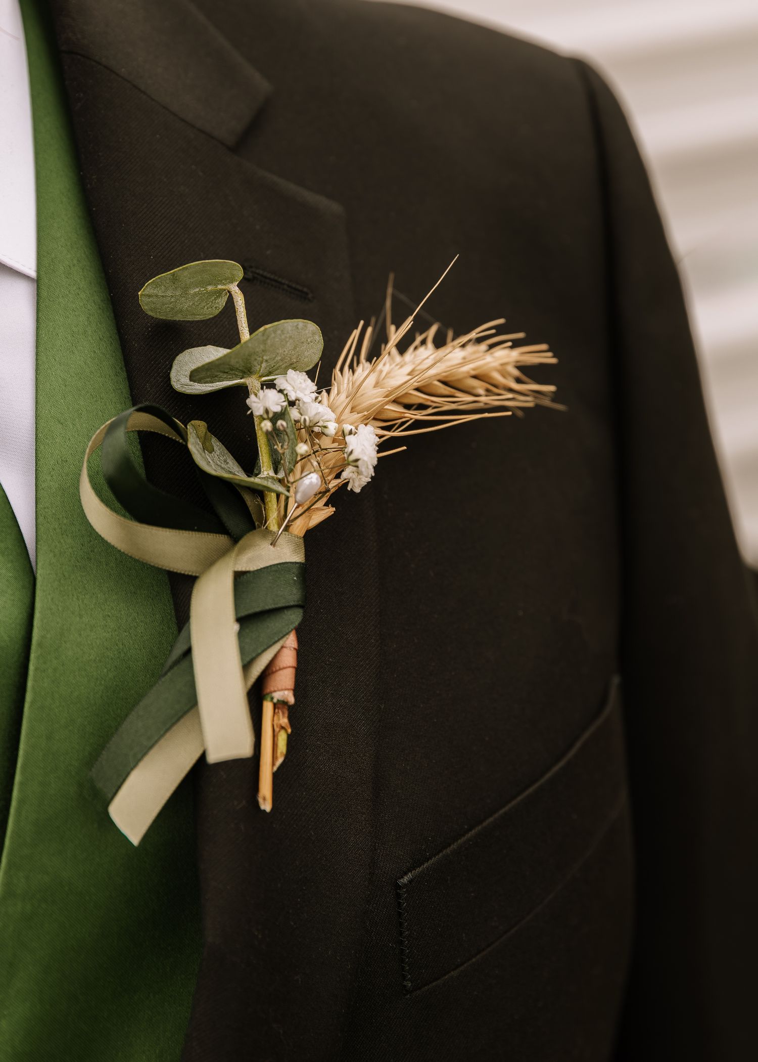 Close-up view of wheat boutonnieres with white ribbons pinned to black suit lapels for a wedding.