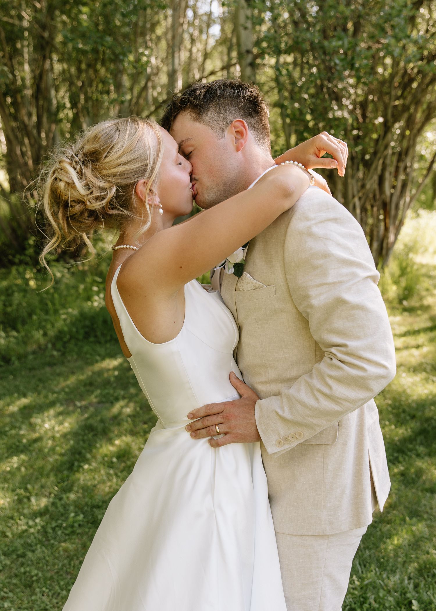 A couple in wedding attire share a romantic kiss in a sunlit wooded setting.