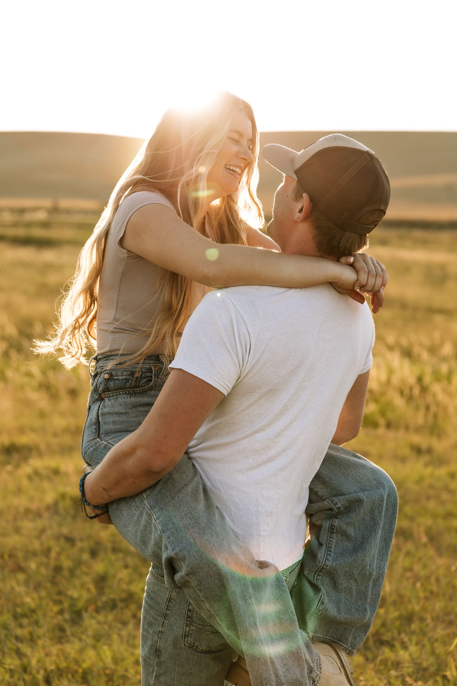 A romantic couple embraces in a sunlit meadow wearing casual denim and white clothing during golden hour.