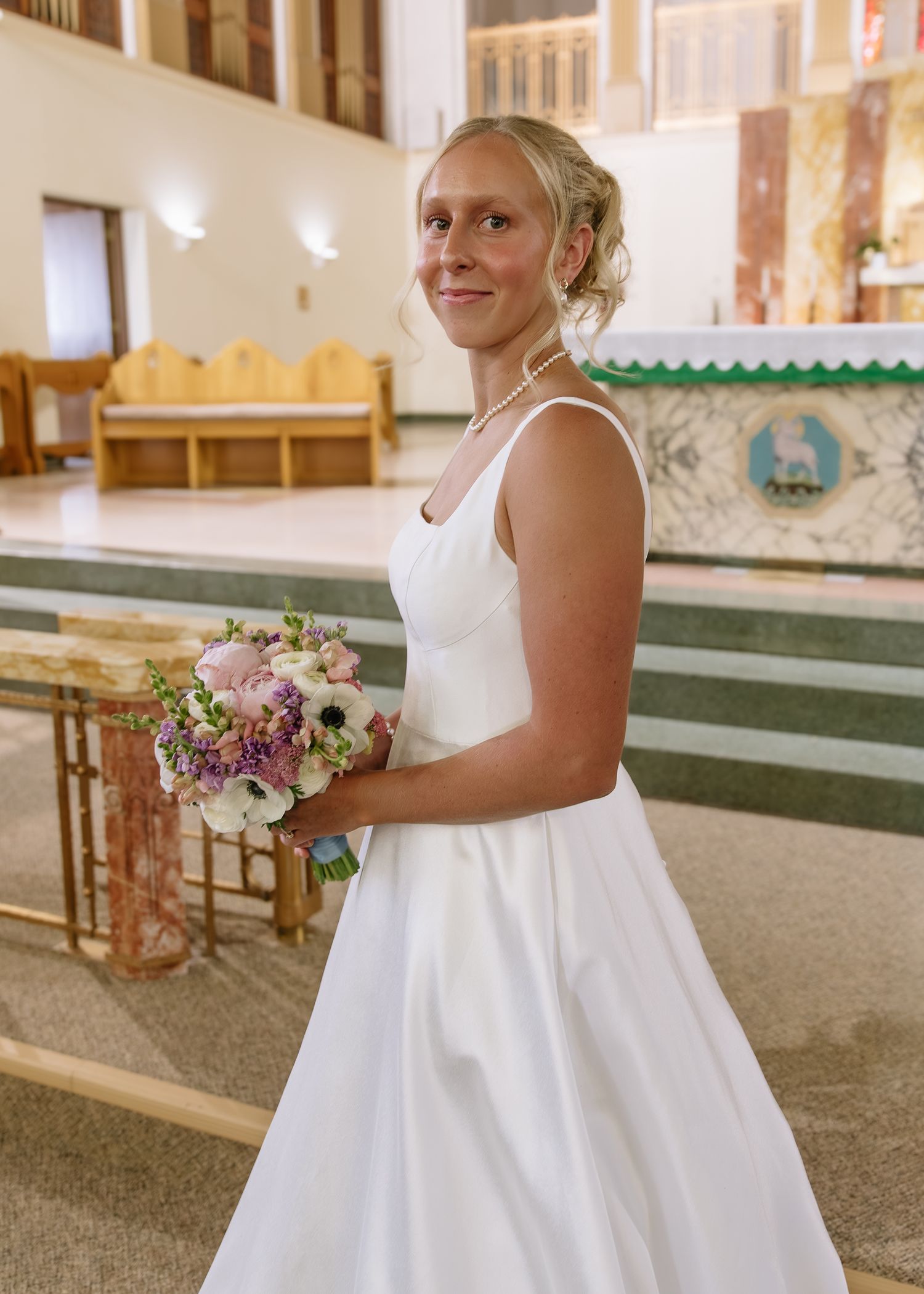 A bride in a classic white wedding dress holds a beautiful bouquet of purple and white flowers inside a church sanctuary.