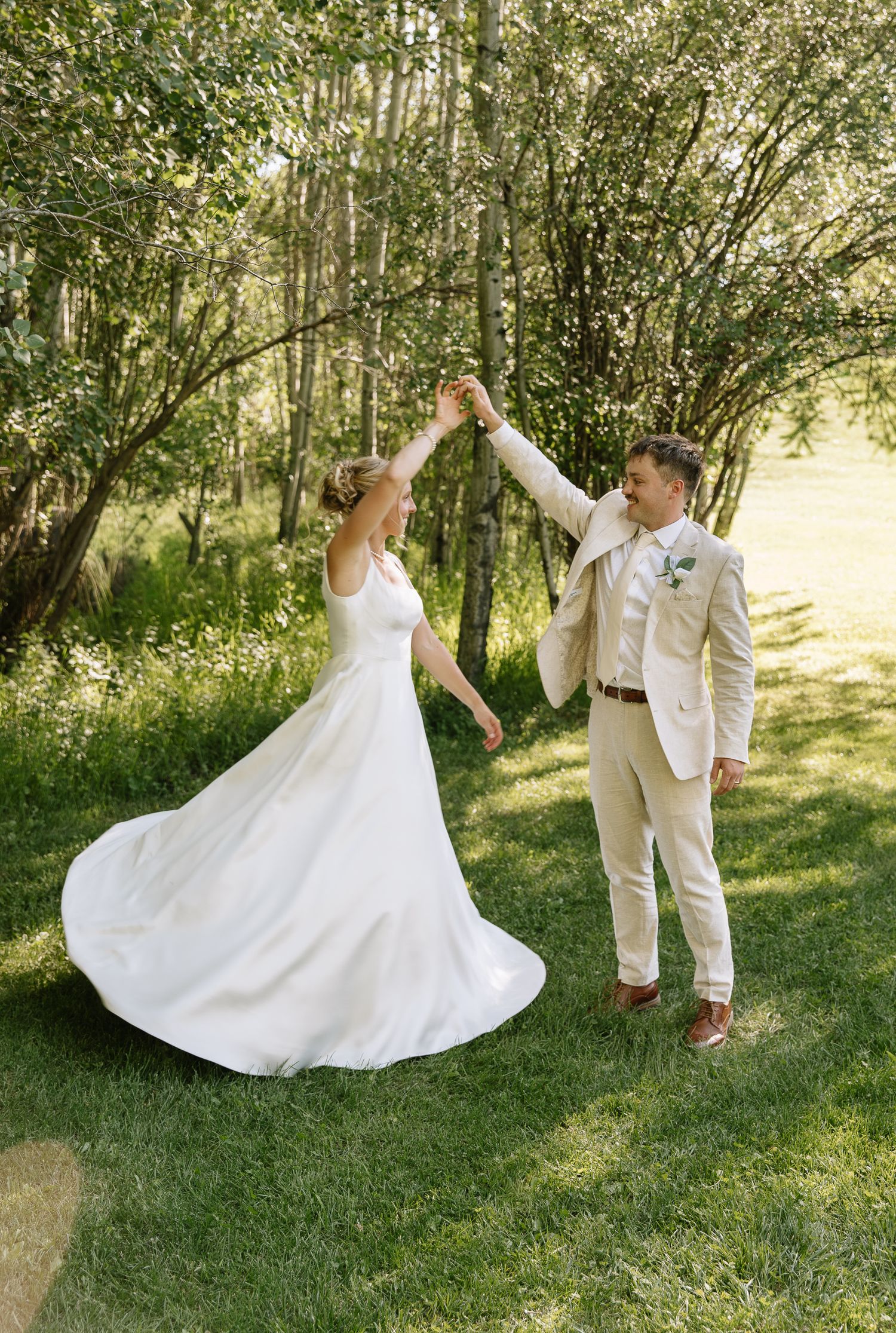 A couple in wedding attire dance together in a sunlit garden setting with trees in the background.