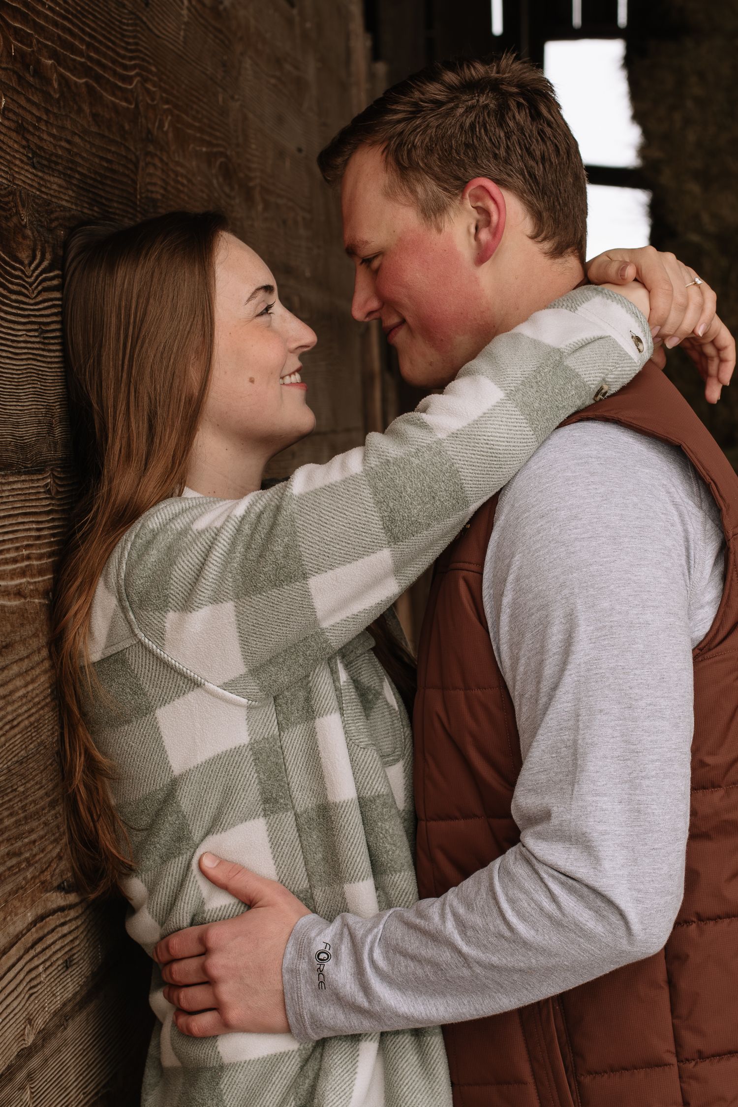 A romantic couple embraces against a rustic wall, with her wearing a plaid sweater and him in a brown vest.