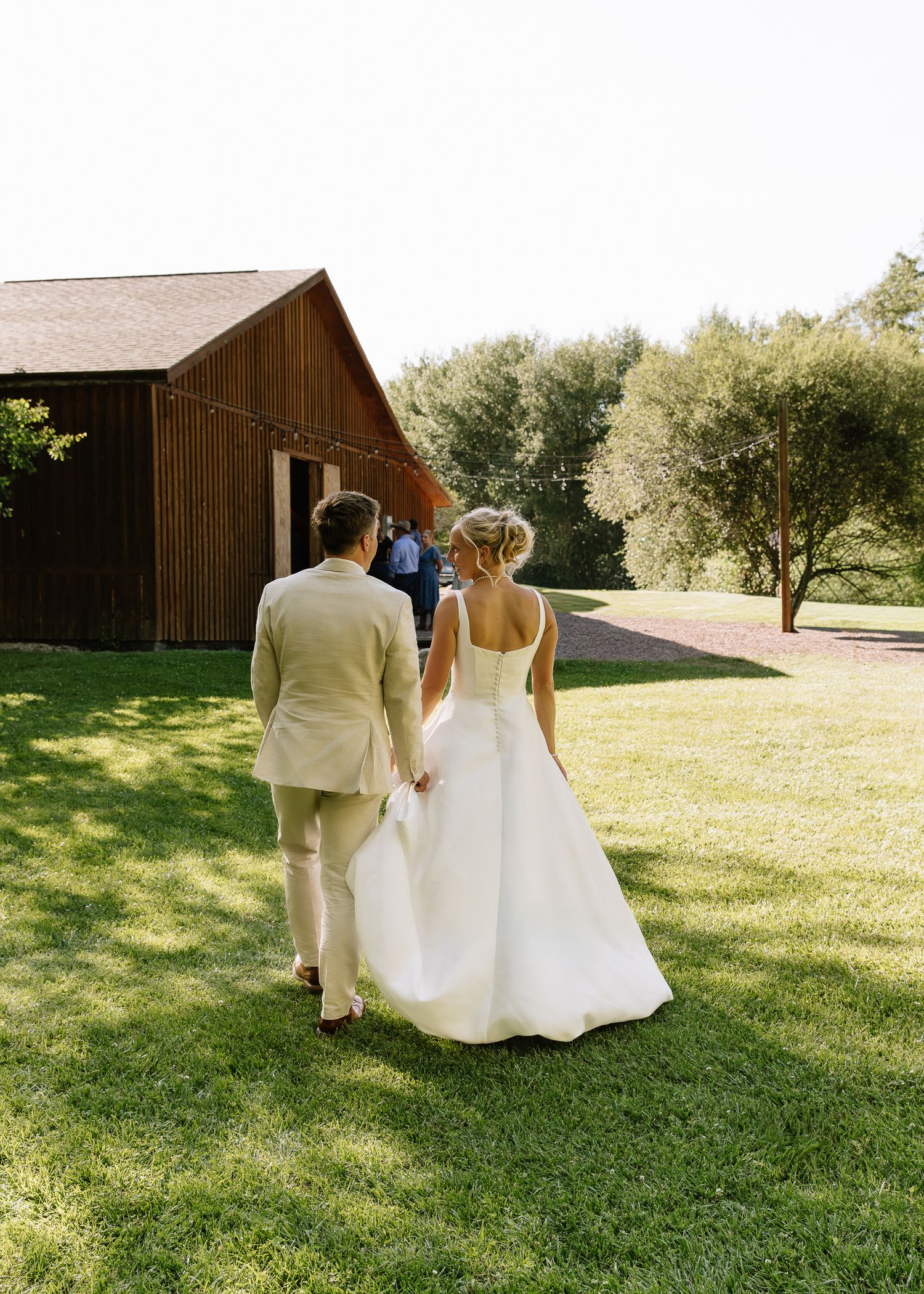 A couple in wedding attire walks hand in hand across a sunlit grassy lawn on their special day.