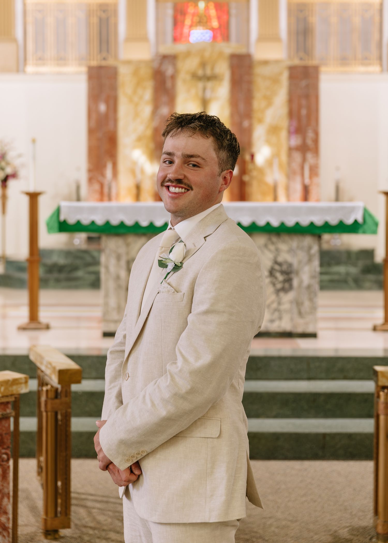 A person in a white suit smiles and poses in a church sanctuary with wooden pews and stained glass windows.