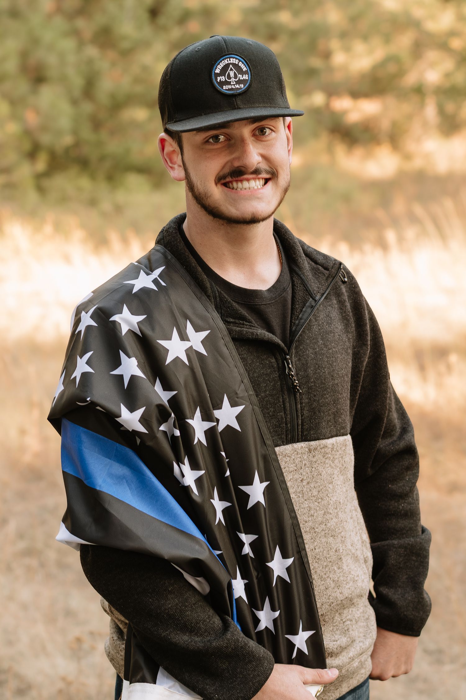 Person wearing a black baseball cap and fleece jacket with a thin blue line flag design smiles outdoors.