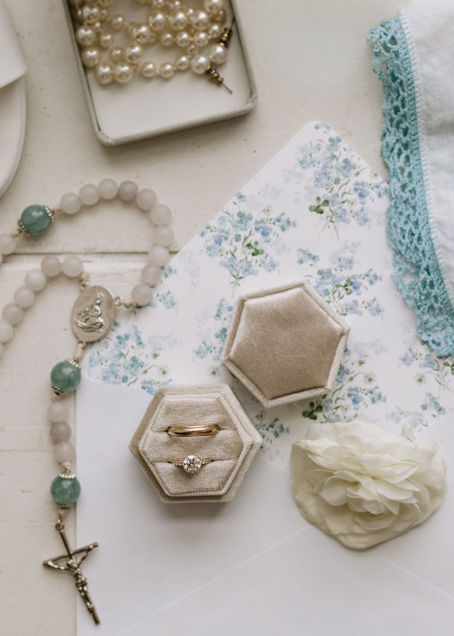 Delicate wedding accessories including pearl necklaces, rings in hexagonal boxes, and blue floral details arranged on white surface.