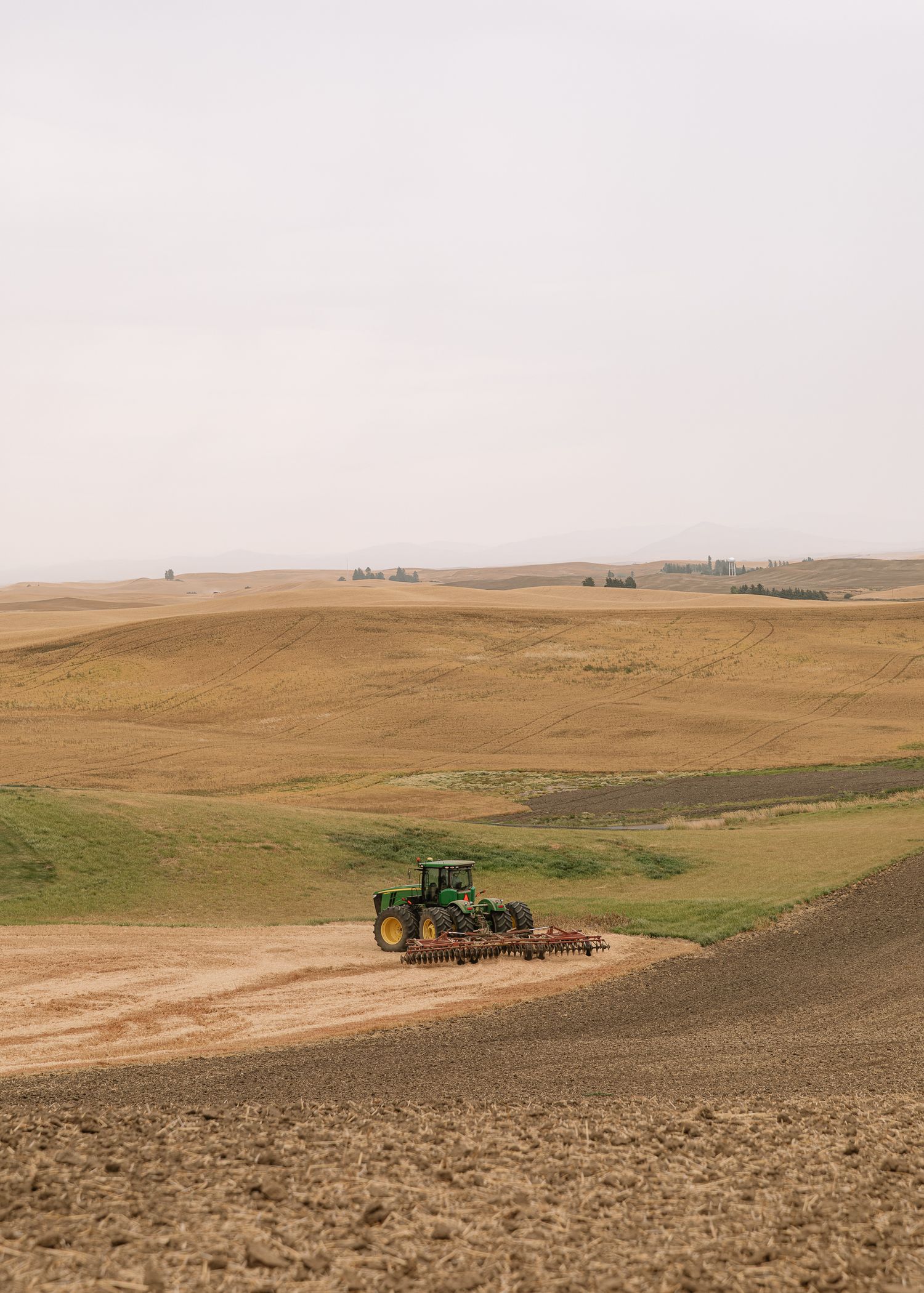 A green John Deere tractor works on rolling hills of farmland with plowed fields stretching to the horizon.