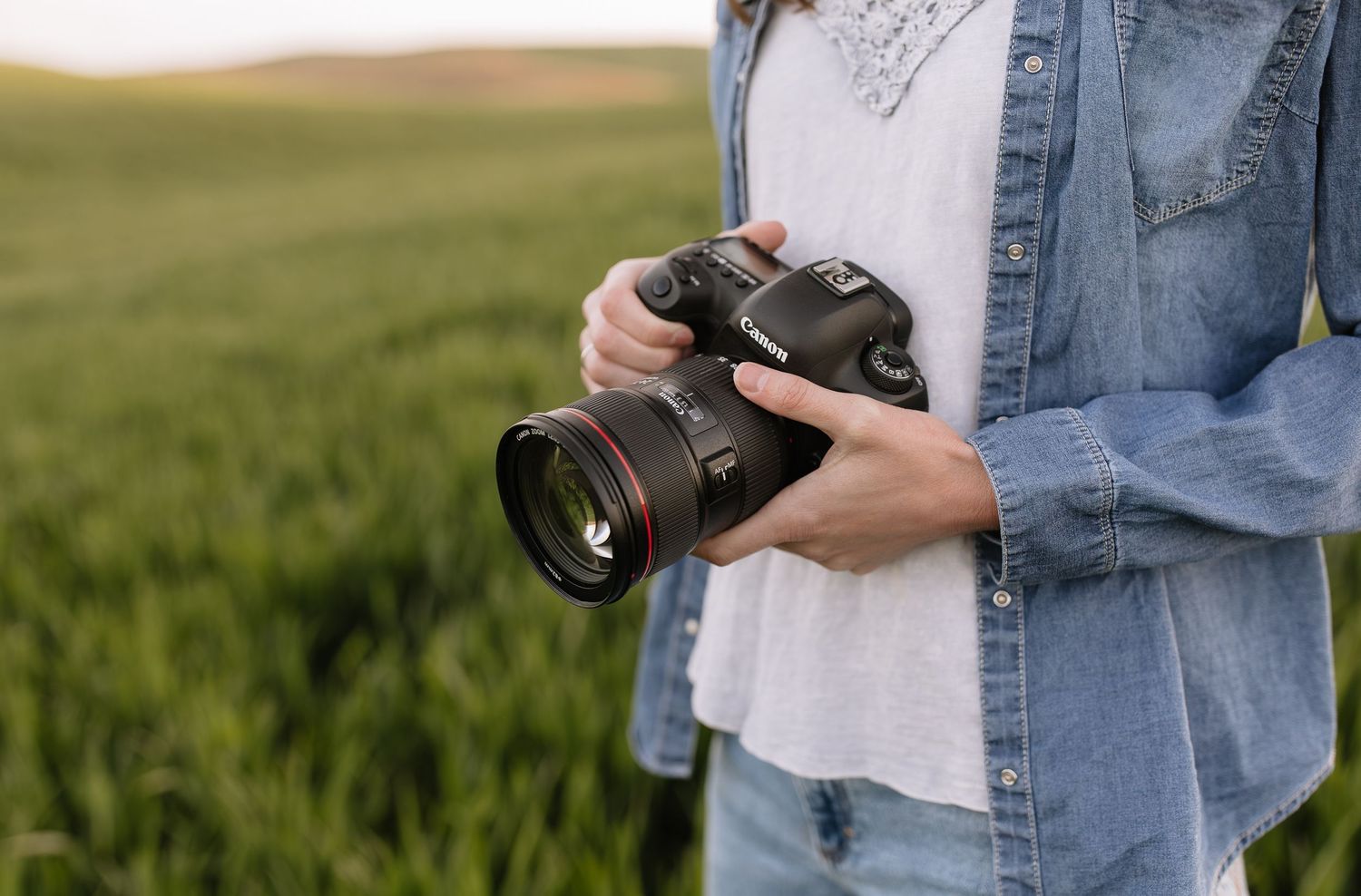 Person holding professional camera while standing in green field wearing denim jacket and white shirt.