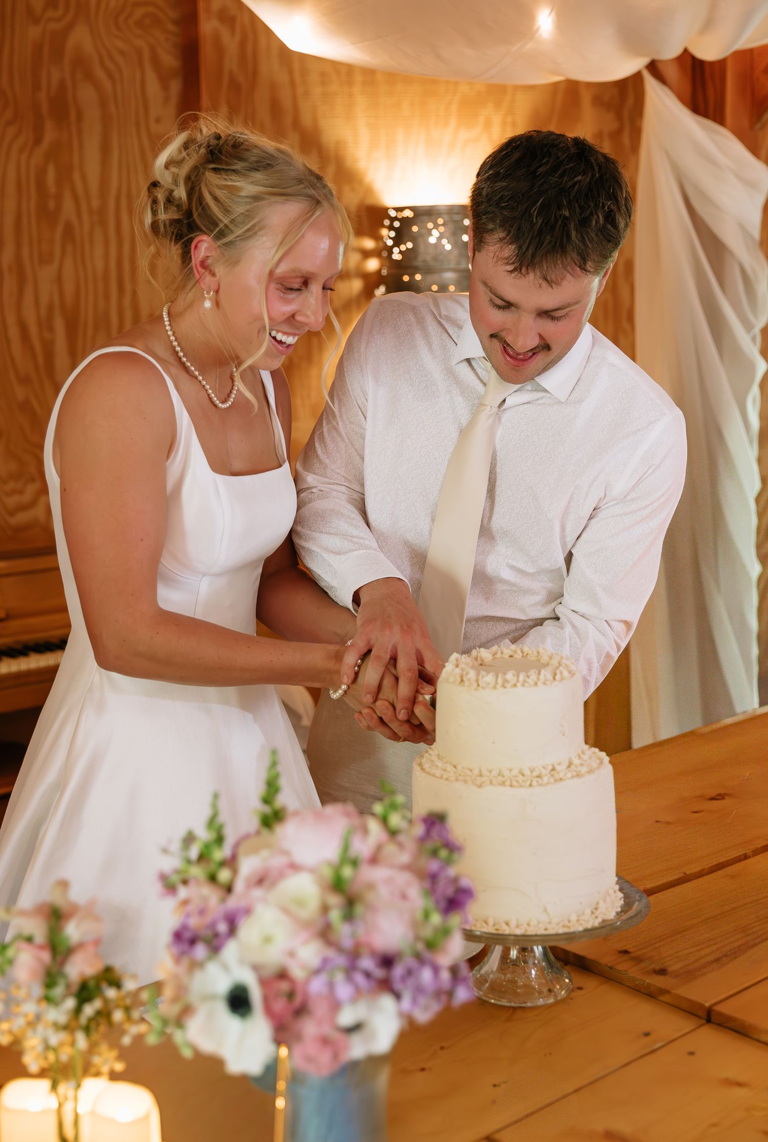 A couple in white attire cuts their two-tiered wedding cake together while floral decorations adorn the wooden table.