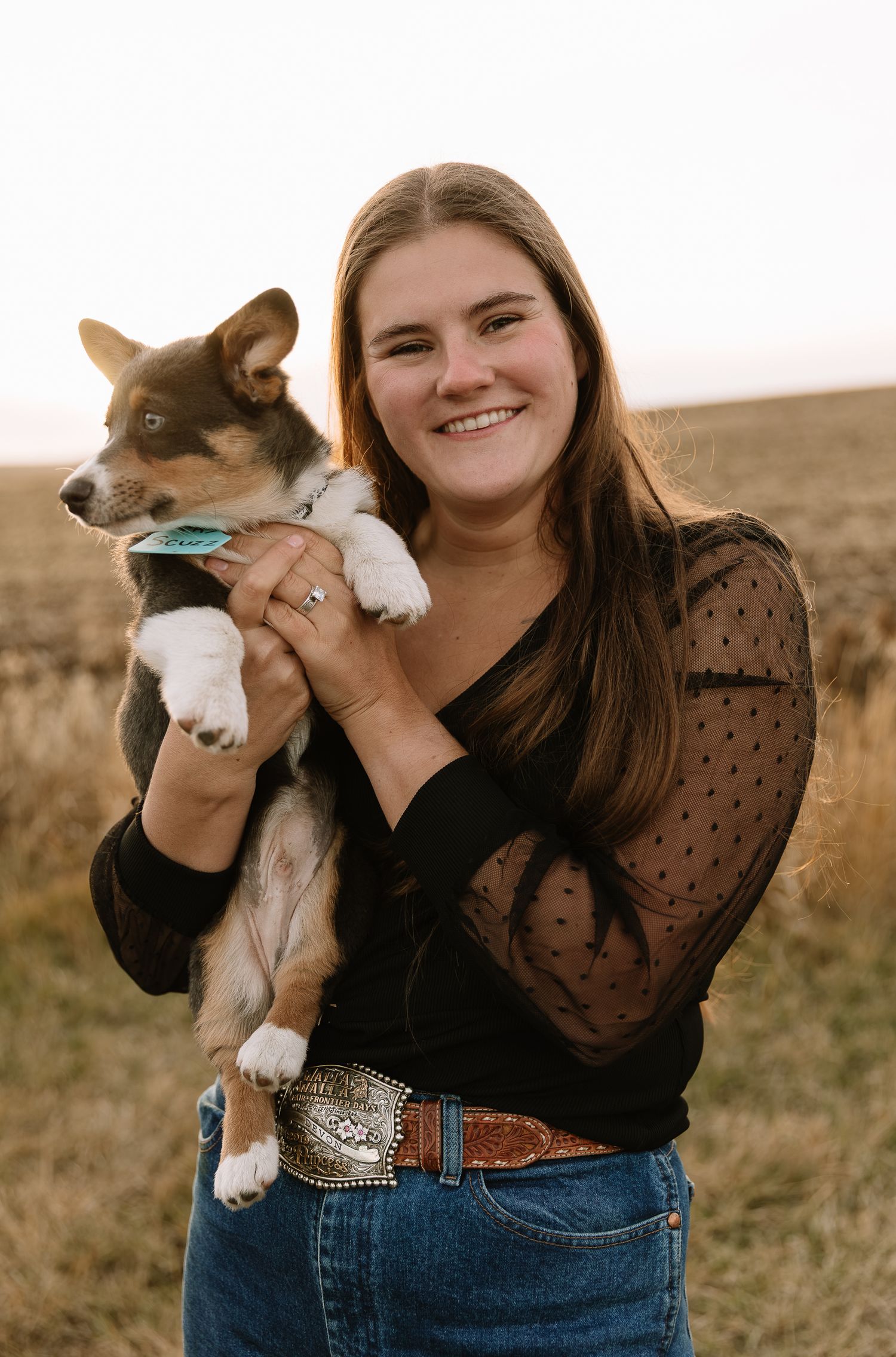 A woman in a polka dot blouse and jeans holds a small brown and white corgi puppy in a field at sunset.