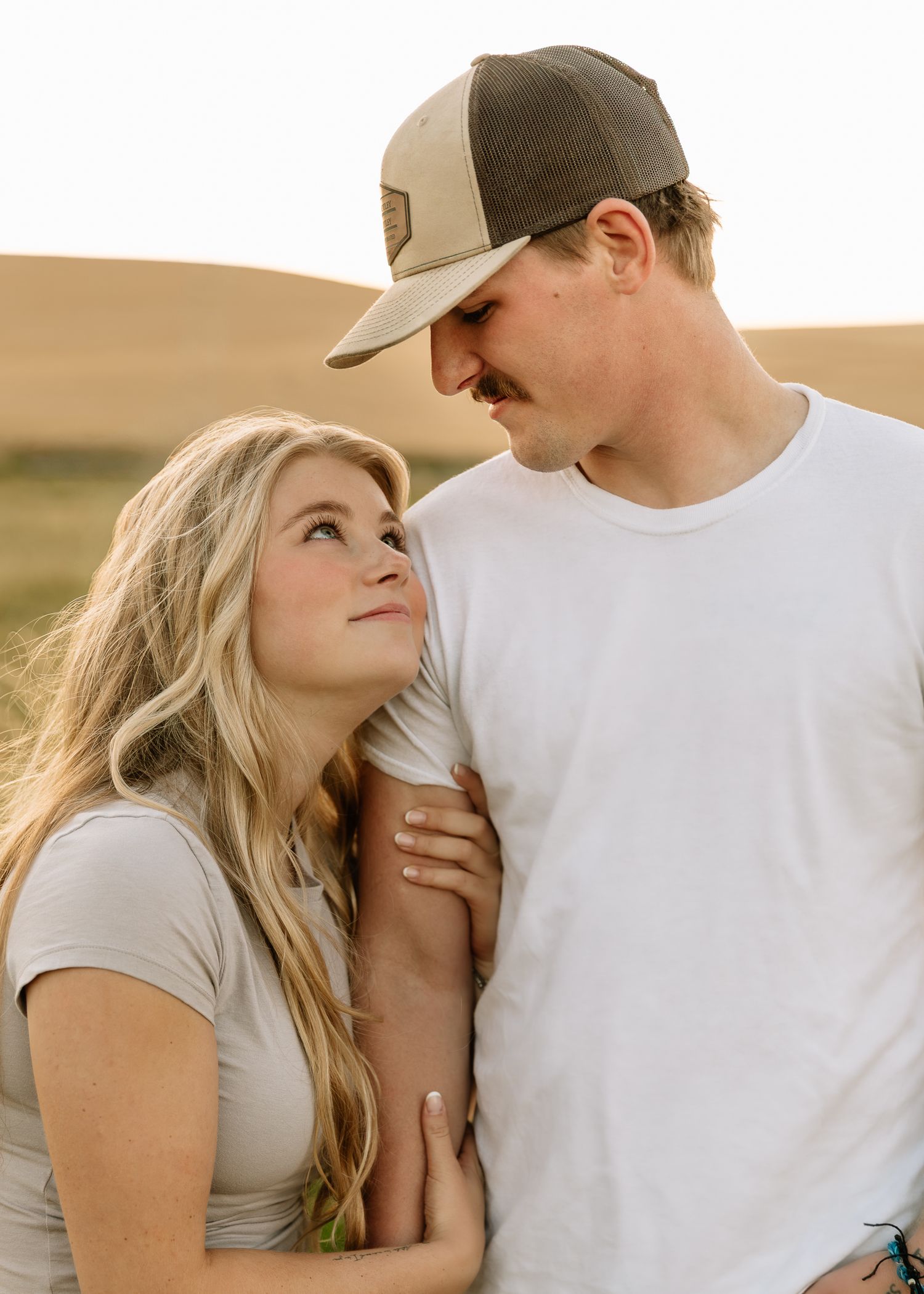 A couple in casual white and gray attire share intimate moments against a warm outdoor backdrop at sunset.