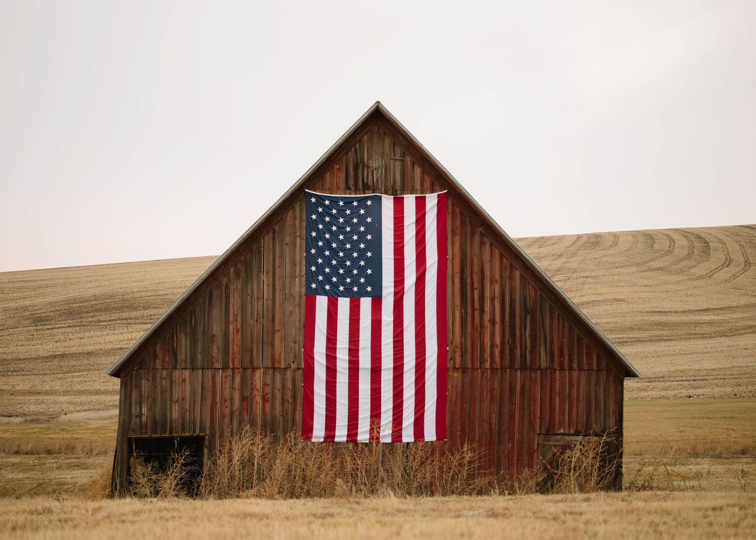A rustic wooden barn with an American flag mounted on its weathered facade stands against rolling hills in the countryside.